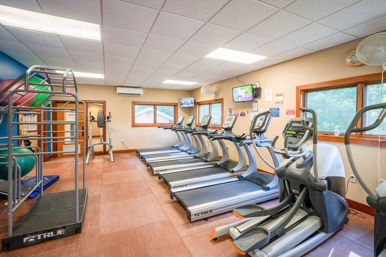 Alt=A gym room with several treadmills, a stationary bike, exercise balls, and a weight rack. The room has large windows, a TV, and a wall-mounted fan. The walls feature health and safety posters.