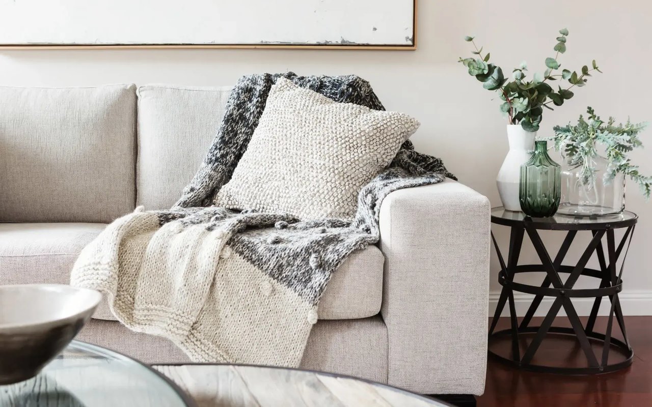 A cozy living room features a gray sofa with a knit blanket, a textured pillow, a glass table, a vase of eucalyptus, and a bowl.