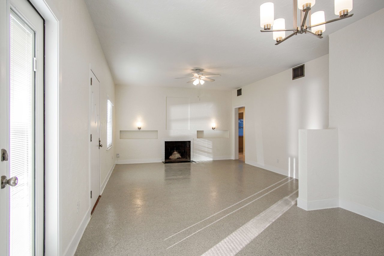 An interior shot of a living room featuring a fireplace, ceiling fan, and tiled flooring. The room is spacious with multiple light fixtures.