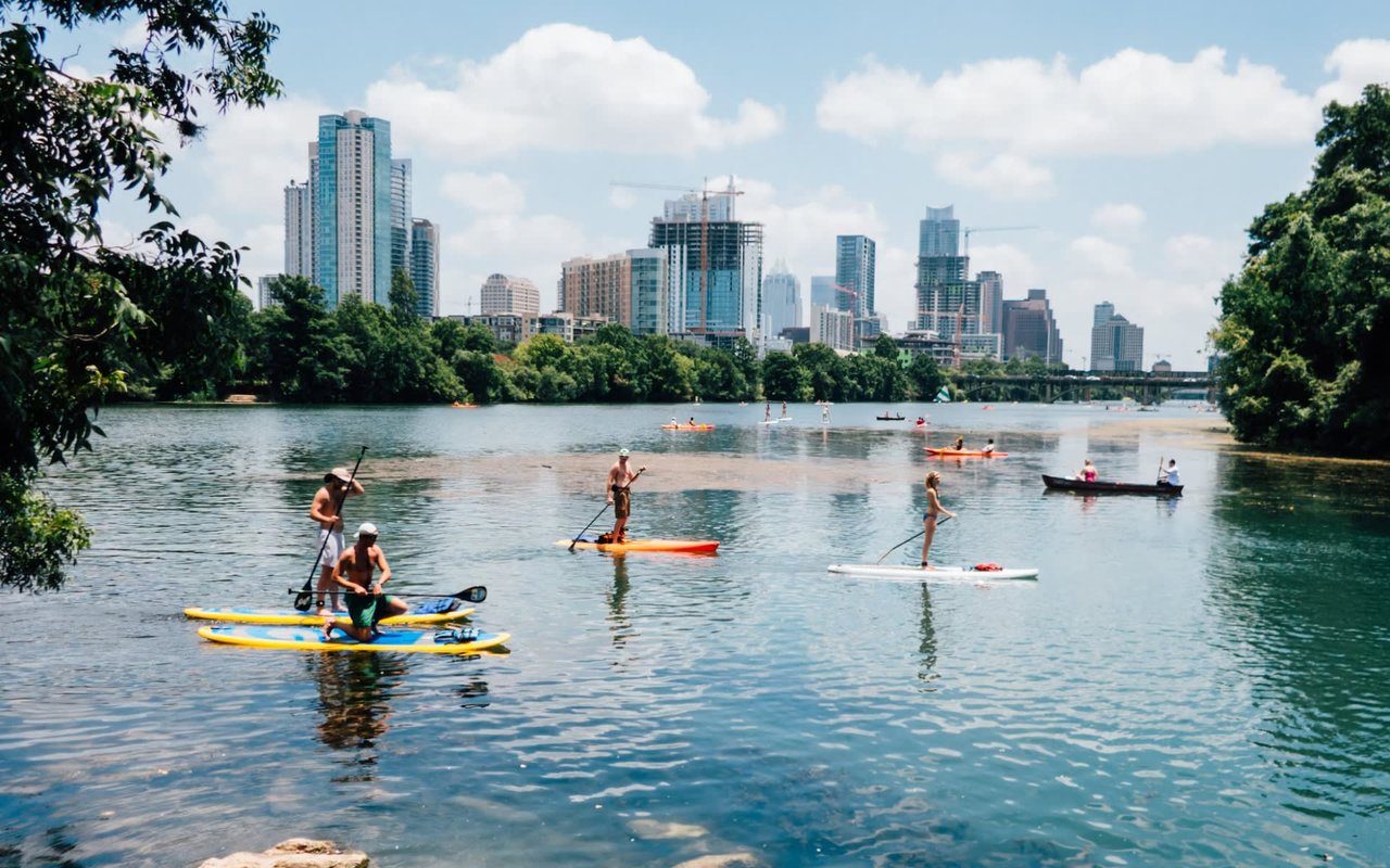 People kayaking on a calm lake surrounded by lush green mountains with great buildings in the background.