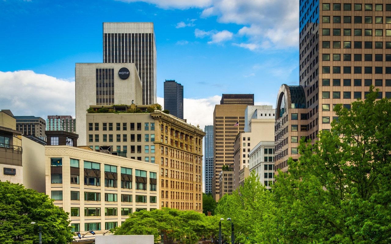 An eye-level view of a bustling city skyline with a mix of modern and classic buildings under a bright blue sky with scattered white clouds.