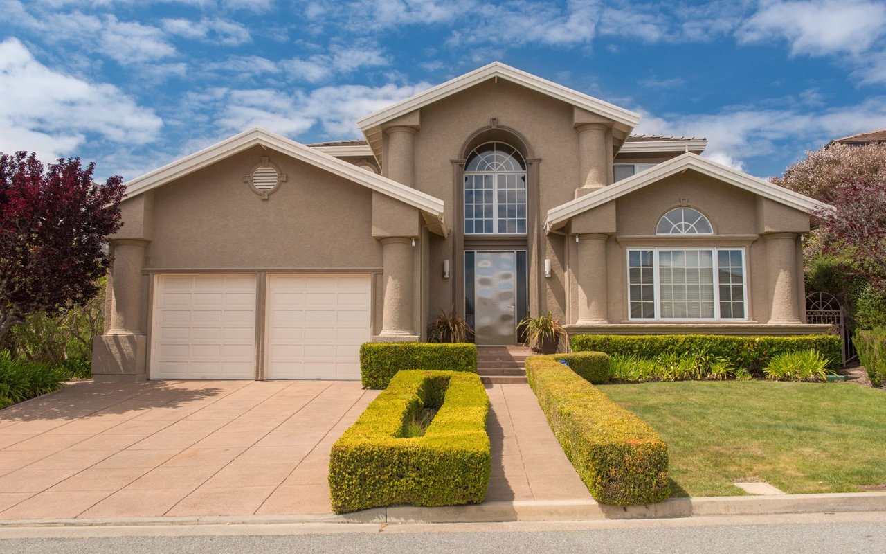 The house has a two-car garage with white doors on the left and multiple large windows with white frames. 