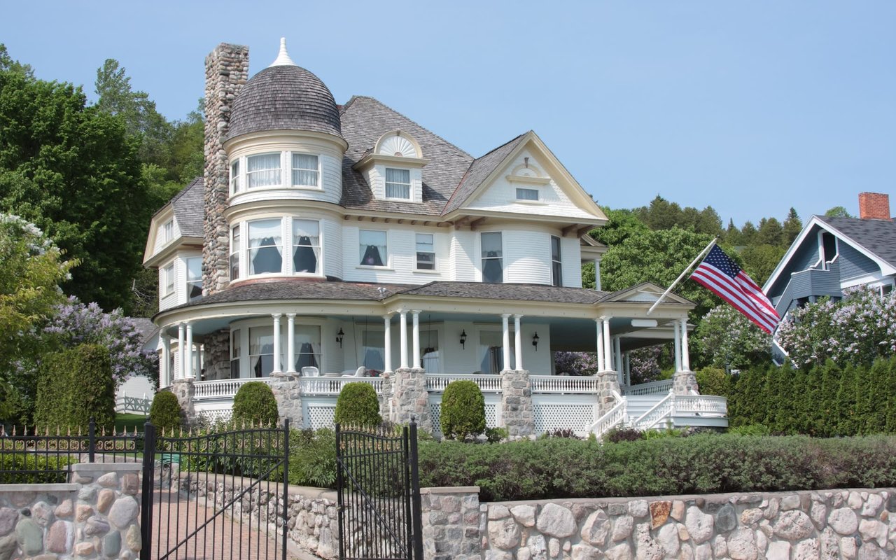 A large white house with a large porch and a stone fence surrounded by trees on a sunny day.