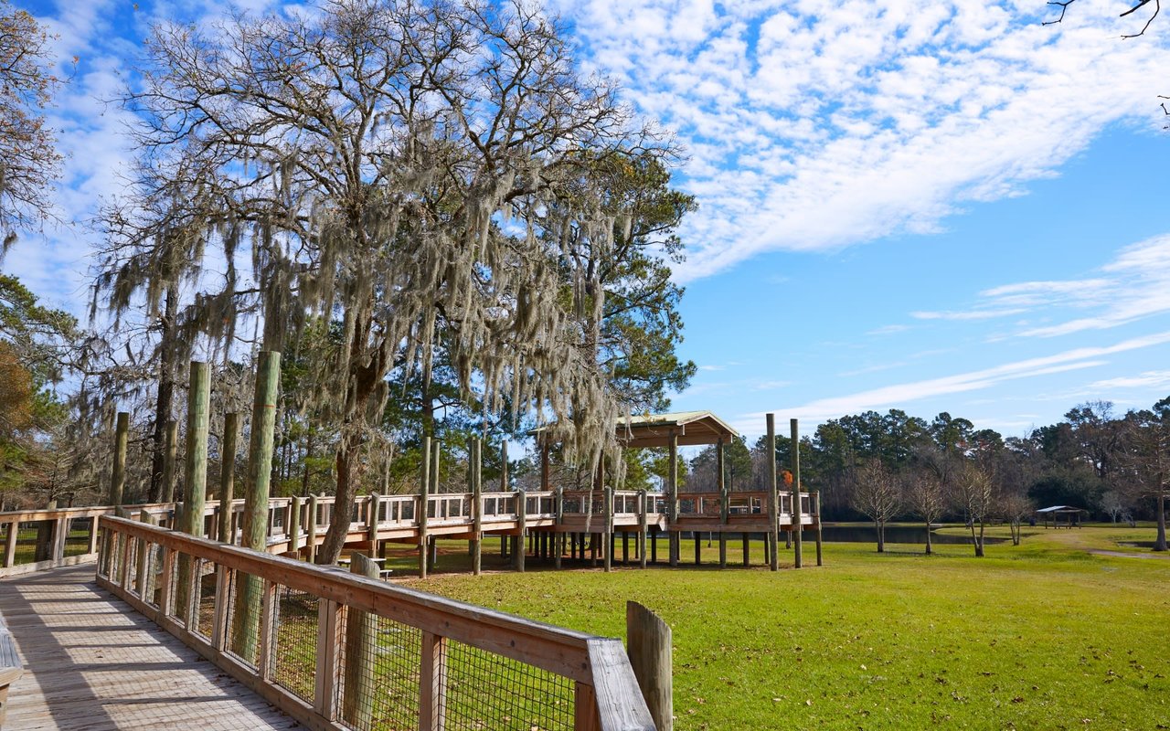 Wooden walkway with railings leading to a hexagonal pavilion in a park, surrounded by grass and mature trees.