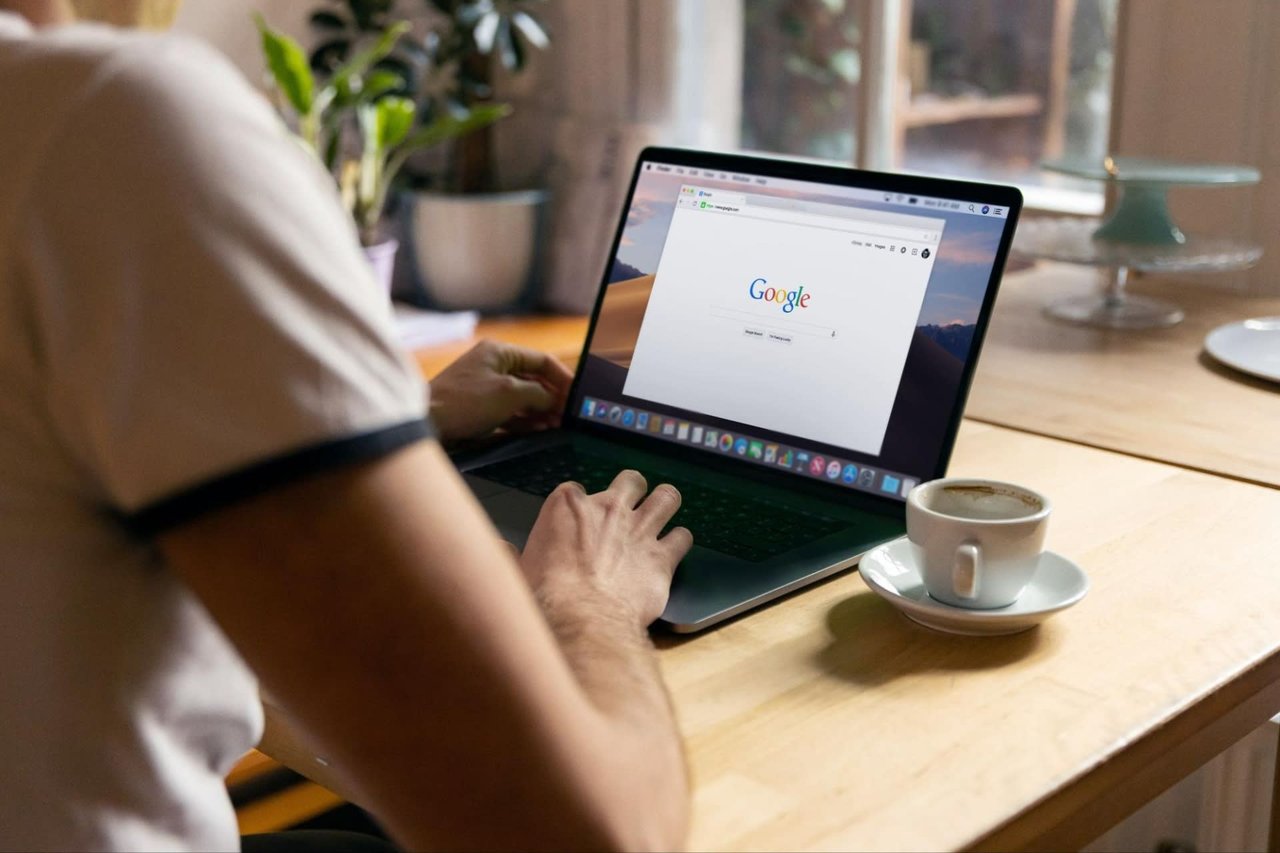 A person sitting at a table using a laptop computer searching on google with a cup of coffee in front of them. 