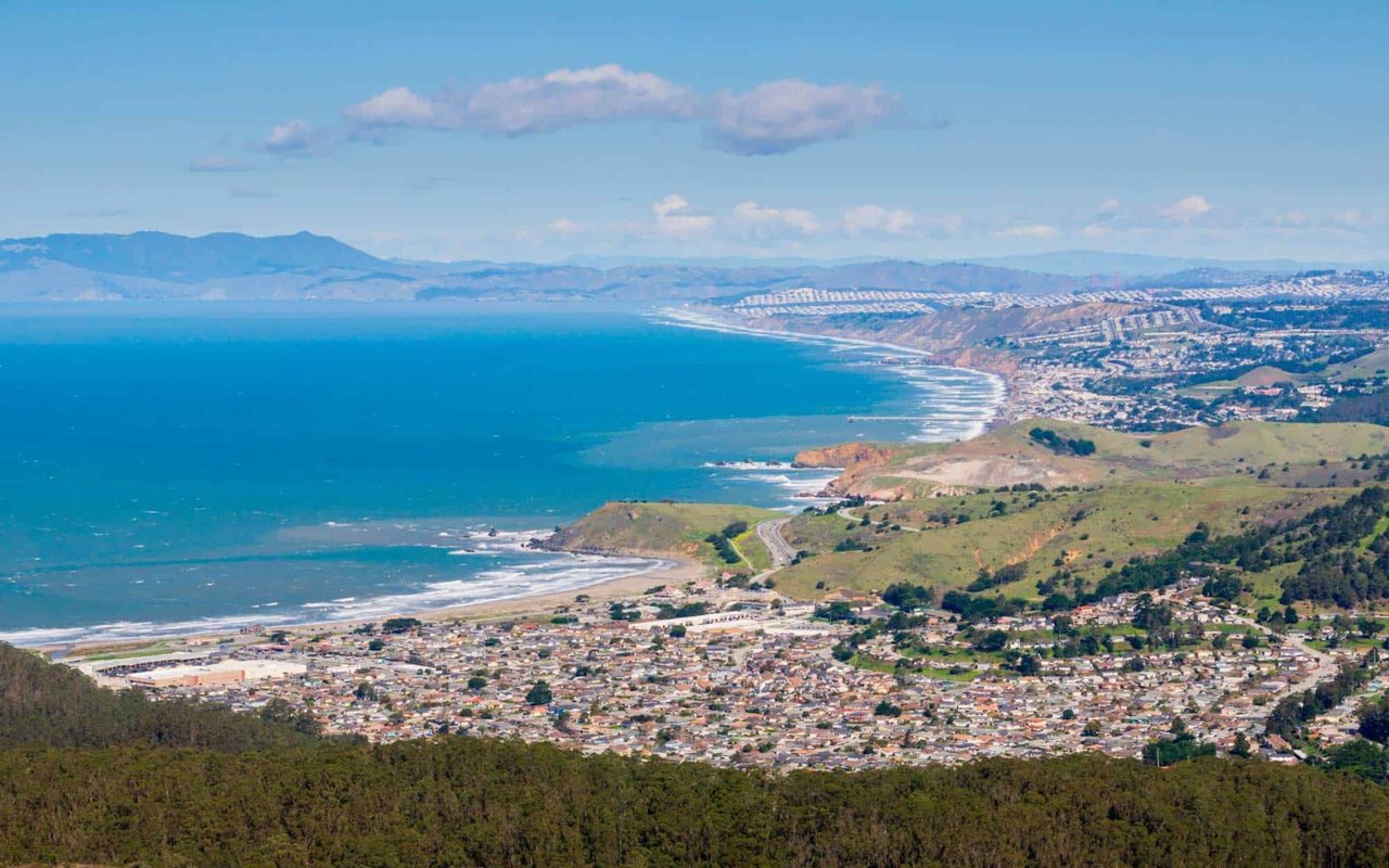 Panoramic aerial view of Pacifica, California, showing coastal homes, rolling green hills, and the Pacific Ocean stretching along the San Mateo County shoreline.