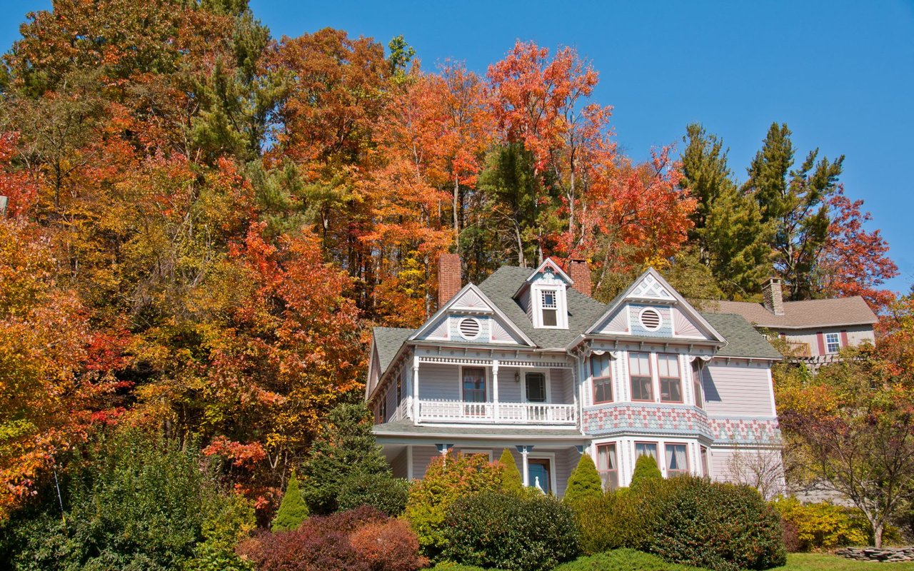 Victorian-style home with white exterior and dark trim, wraparound porch, pitched roof, chimneys, balcony, and front yard.