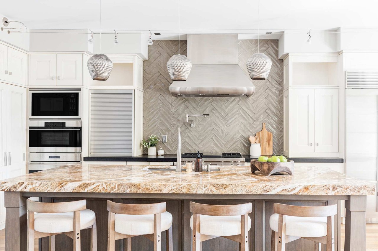 Kitchen with marble island, white cabinets, chevron backsplash, pendant lights, and cream barstools.