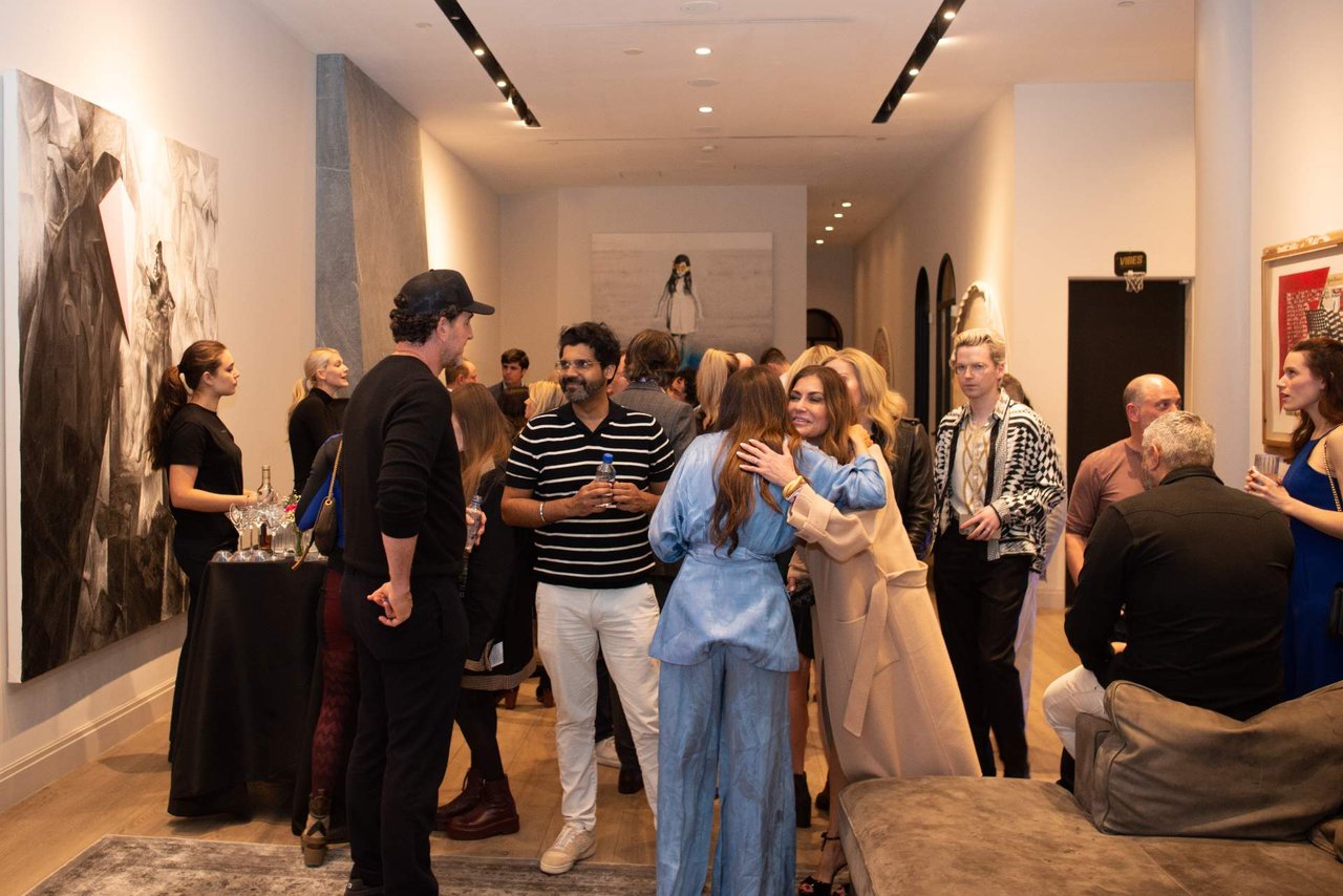 Guests mingle among gallery artwork, including a woman in blue embracing another guest.
