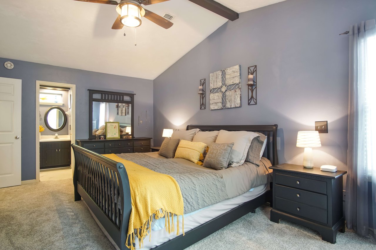 A bedroom with a blue wall color scheme. The room has a large bed, nightstands with lamps, and a ceiling fan. The walls are painted blue, and the room is well-lit.