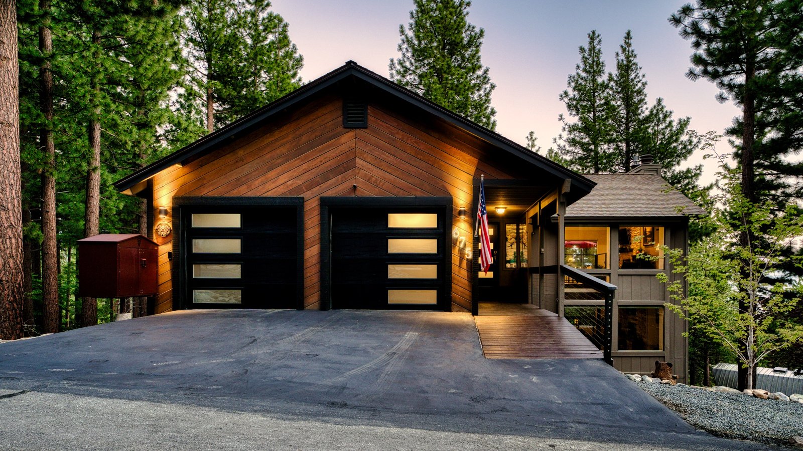 Cedar-sided home with two black glass-paneled garage doors, a covered entry and an American flag, lit at dusk among tall pines at 675 Tyner Way in Lower Tyner, Incline Village