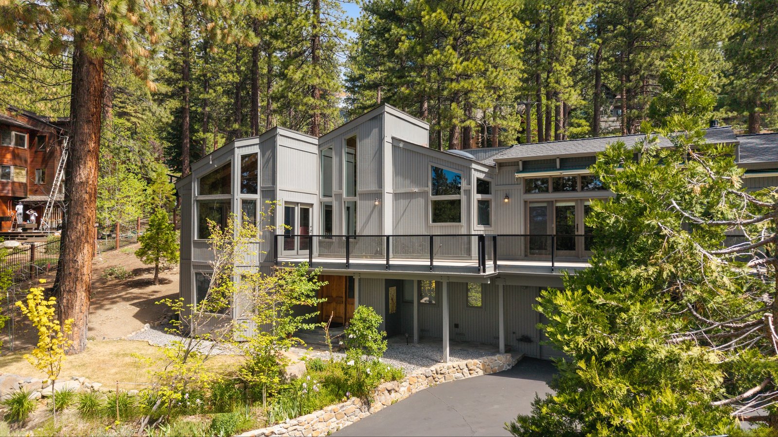 Elevated view of a gray vertical-sided contemporary home with a black-railed deck, a stone-edged driveway and young aspens in a pine forest at 520 Lodgepole Drive, Ponderosa, Incline Village