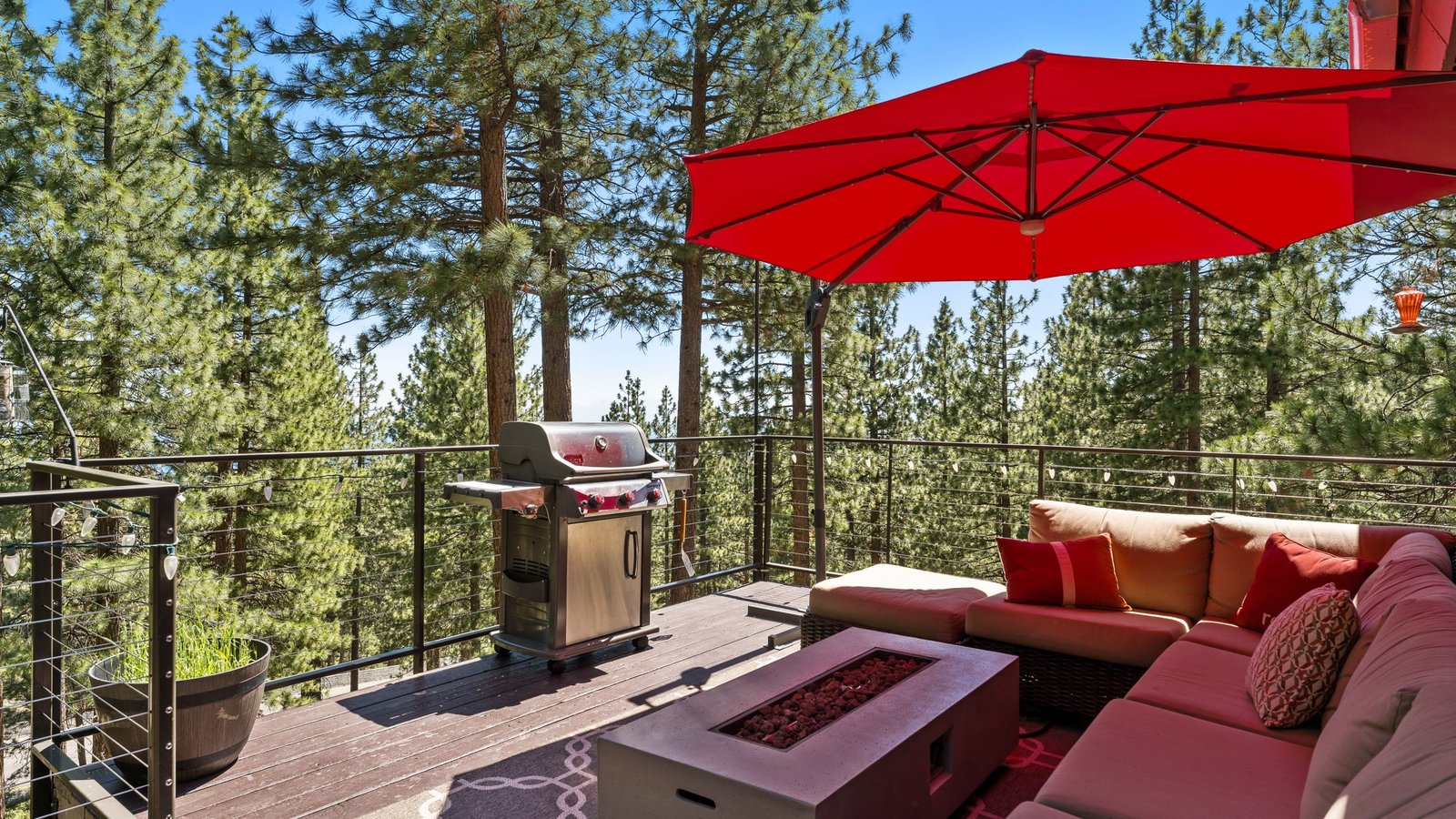 Wood deck with a cable railing, a red market umbrella, a gas fire table, a grill and a sectional sofa, surrounded by tall pines at 675 Tyner Way in Lower Tyner, Incline Village