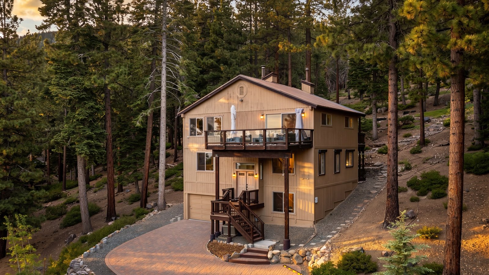 Three-story wood-sided home with an upper deck, lit windows and a paver driveway on a wooded slope at golden hour, 671 Saddlehorn Drive in Lower Tyner, Incline Village