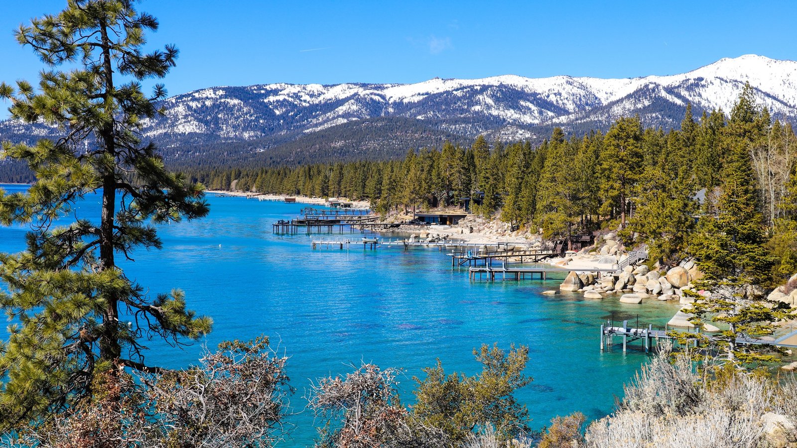 Turquoise Lake Tahoe water, piers and granite boulders below snowcapped peaks near Incline Village