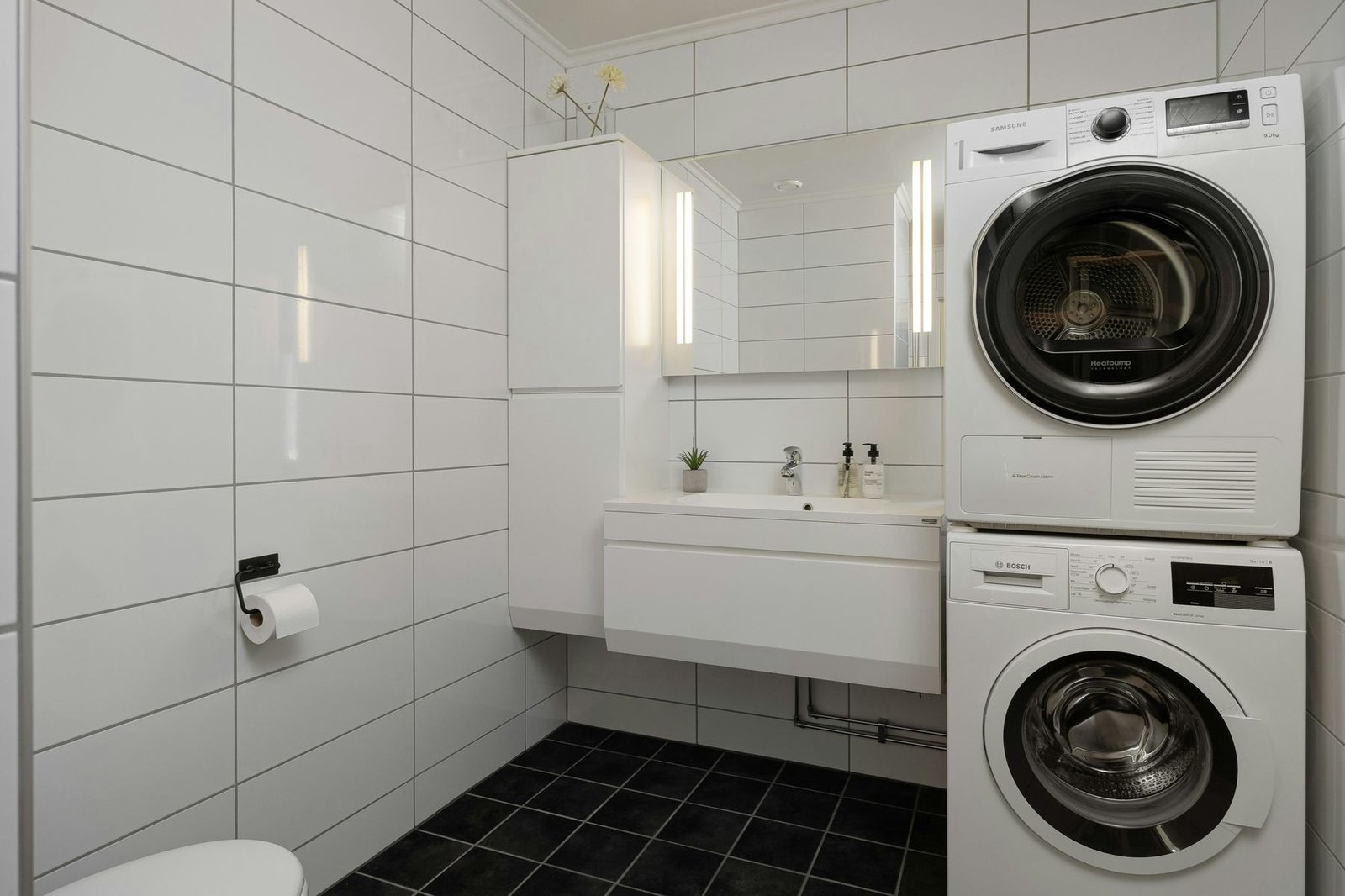Stacked heat-pump dryer and washer in a white tiled bathroom.