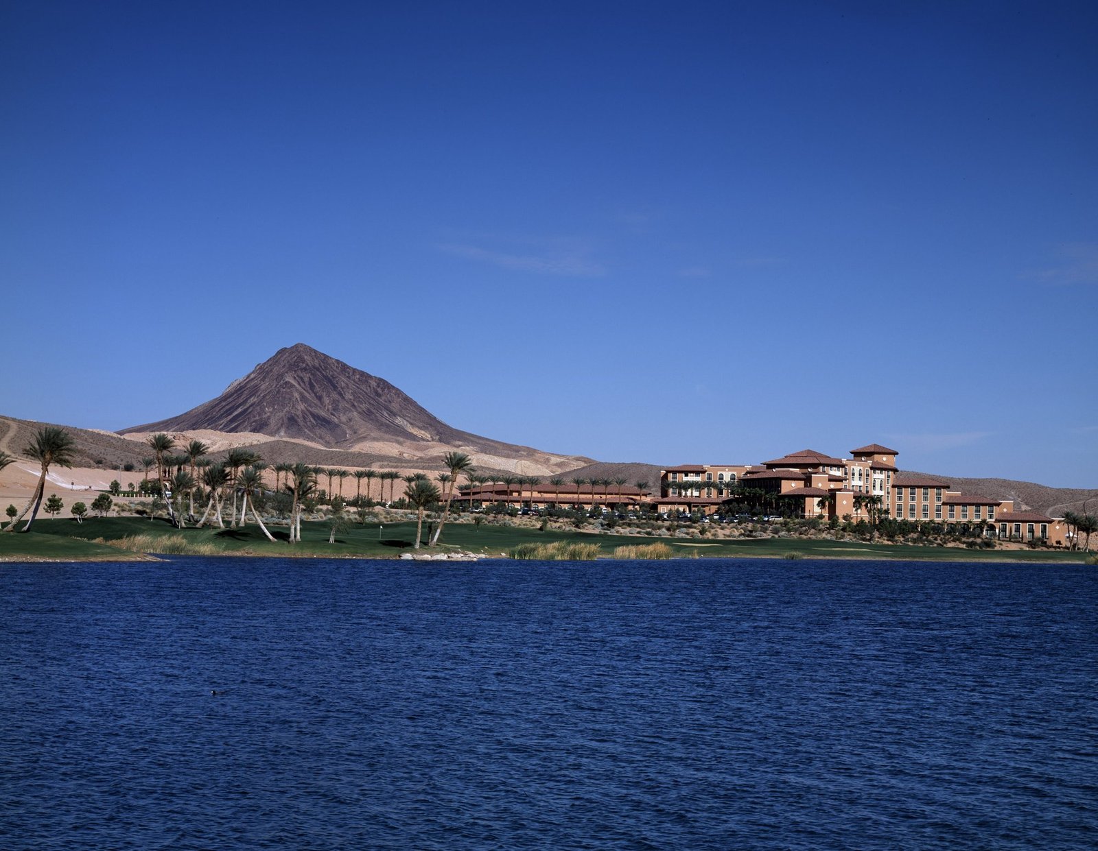 Blue water of Lake Las Vegas in front of palm trees, a green lawn and a Mediterranean-style resort, with a desert peak under a clear sky