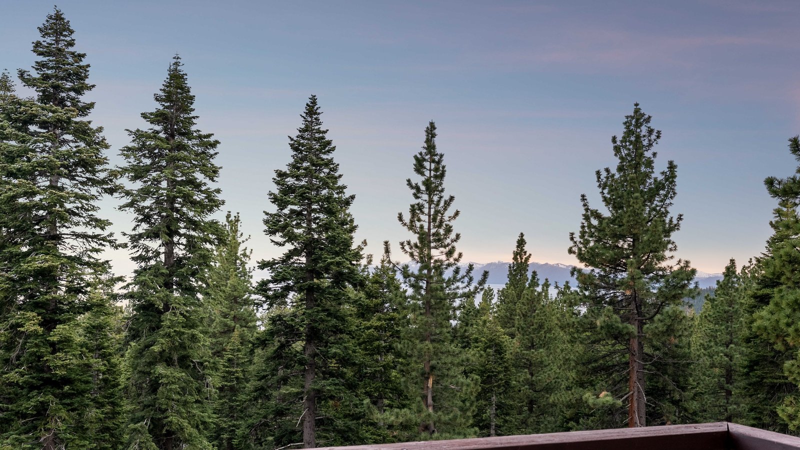 Tall pines at dusk with Lake Tahoe and the snow-dusted peaks of the far shore on the horizon, seen over a deck railing at 671 Saddlehorn Drive in Lower Tyner, Incline Village