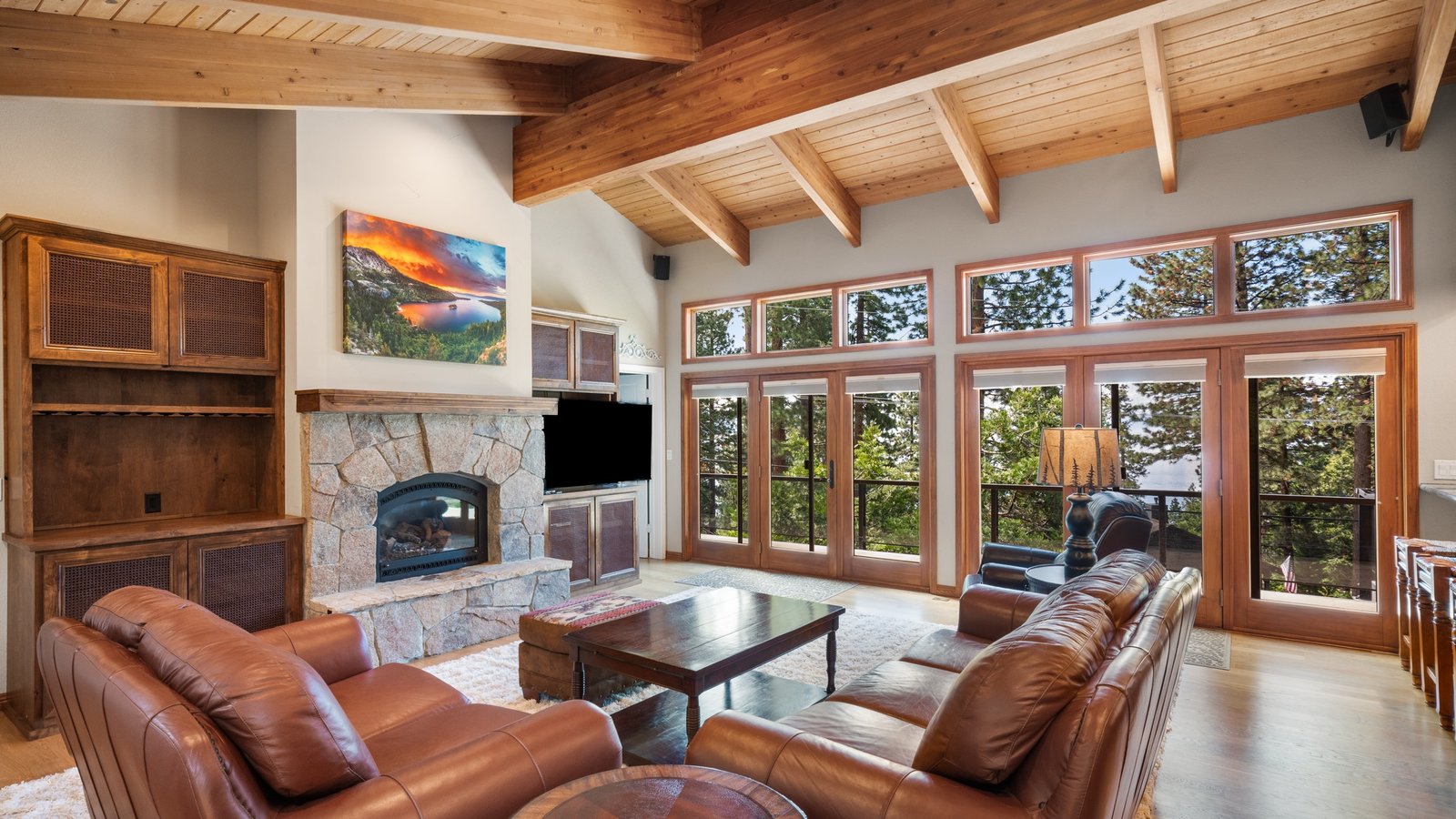 Great room with a vaulted wood-plank ceiling and exposed beams, a stone fireplace, leather sofas and a wall of glass doors opening to a deck in the pines at 520 Lodgepole Drive, Ponderosa, Incline Village