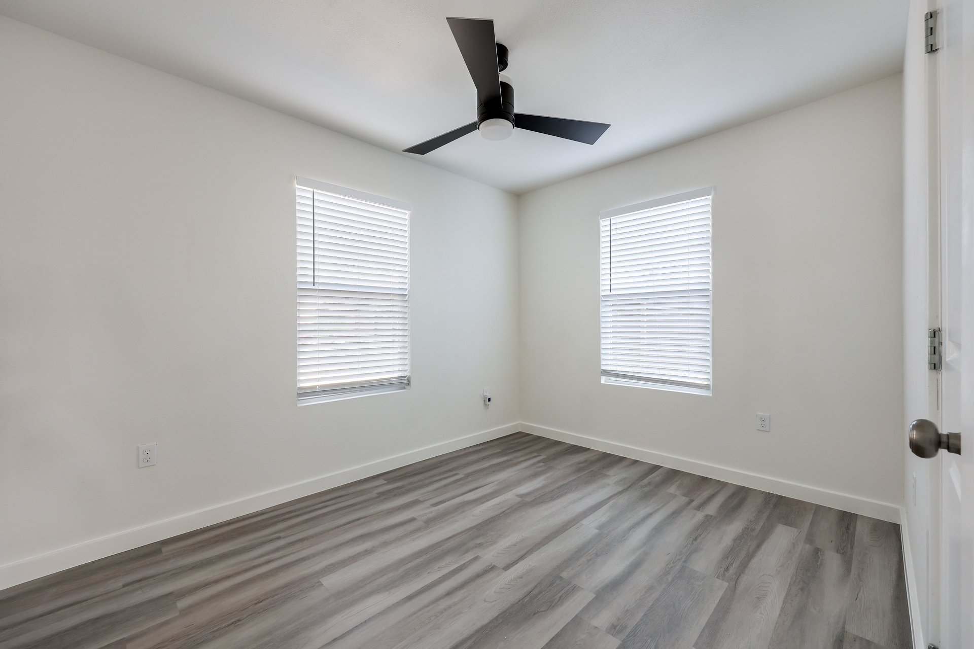 Empty bedroom with gray plank flooring, two windows with blinds, and a black ceiling fan.
