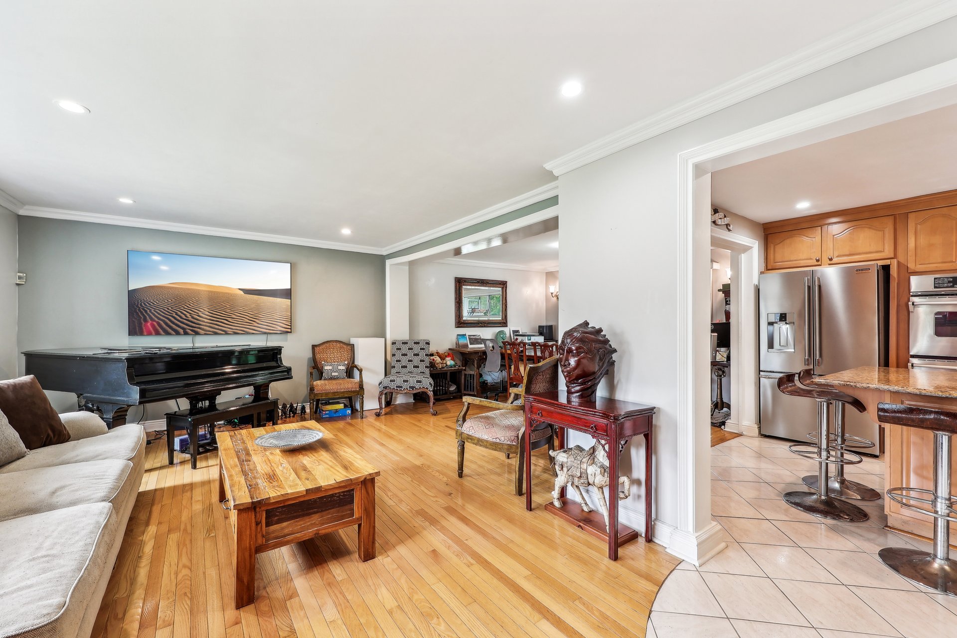 Living room with a grand piano and hardwood floors, opening to a kitchen with wood cabinets.