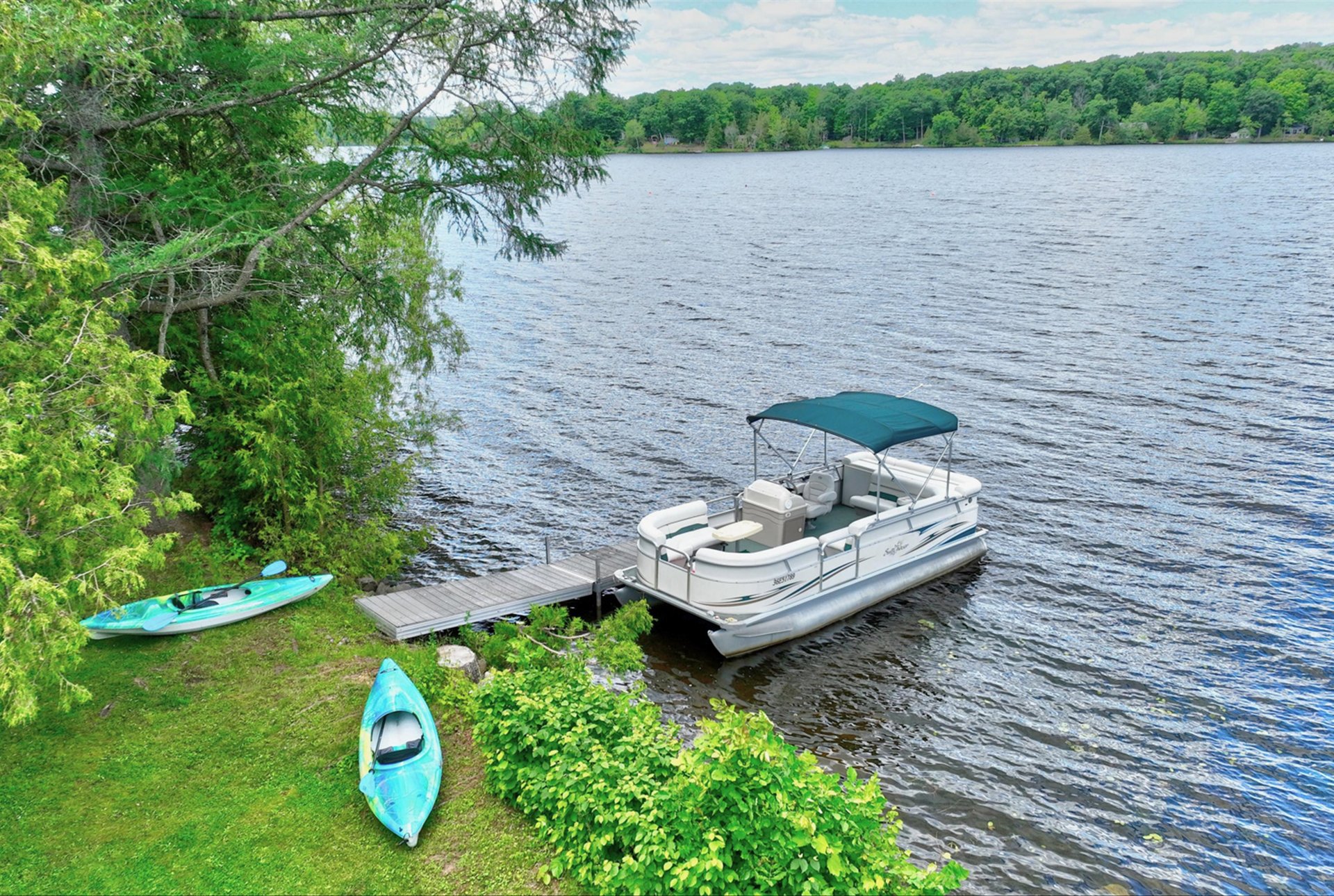 Wooded waterfront with a dock, pontoon boat, and two kayaks beside the lake.