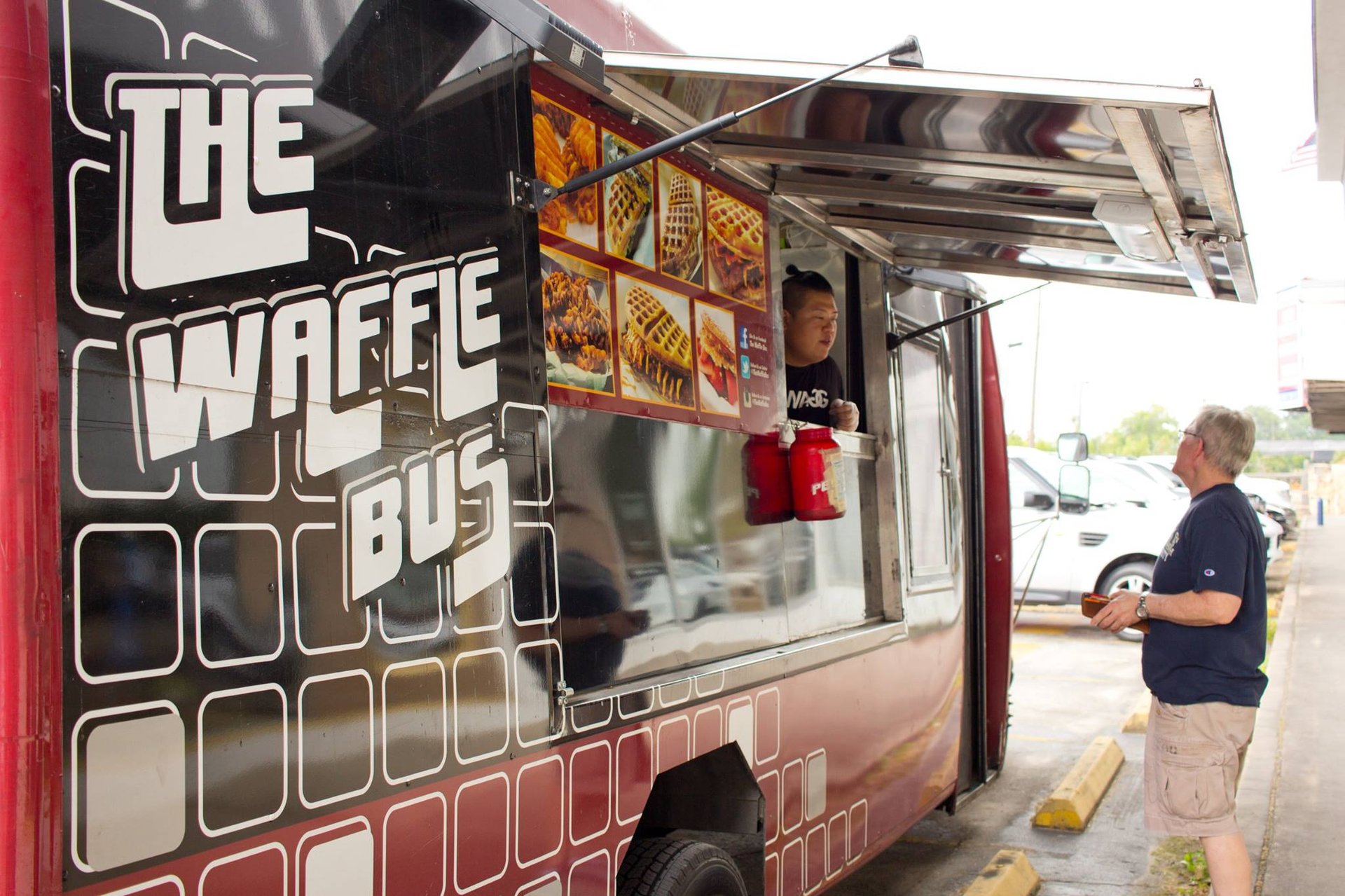 Customer ordering at The Waffle Bus, a red food truck displaying photos of waffles.