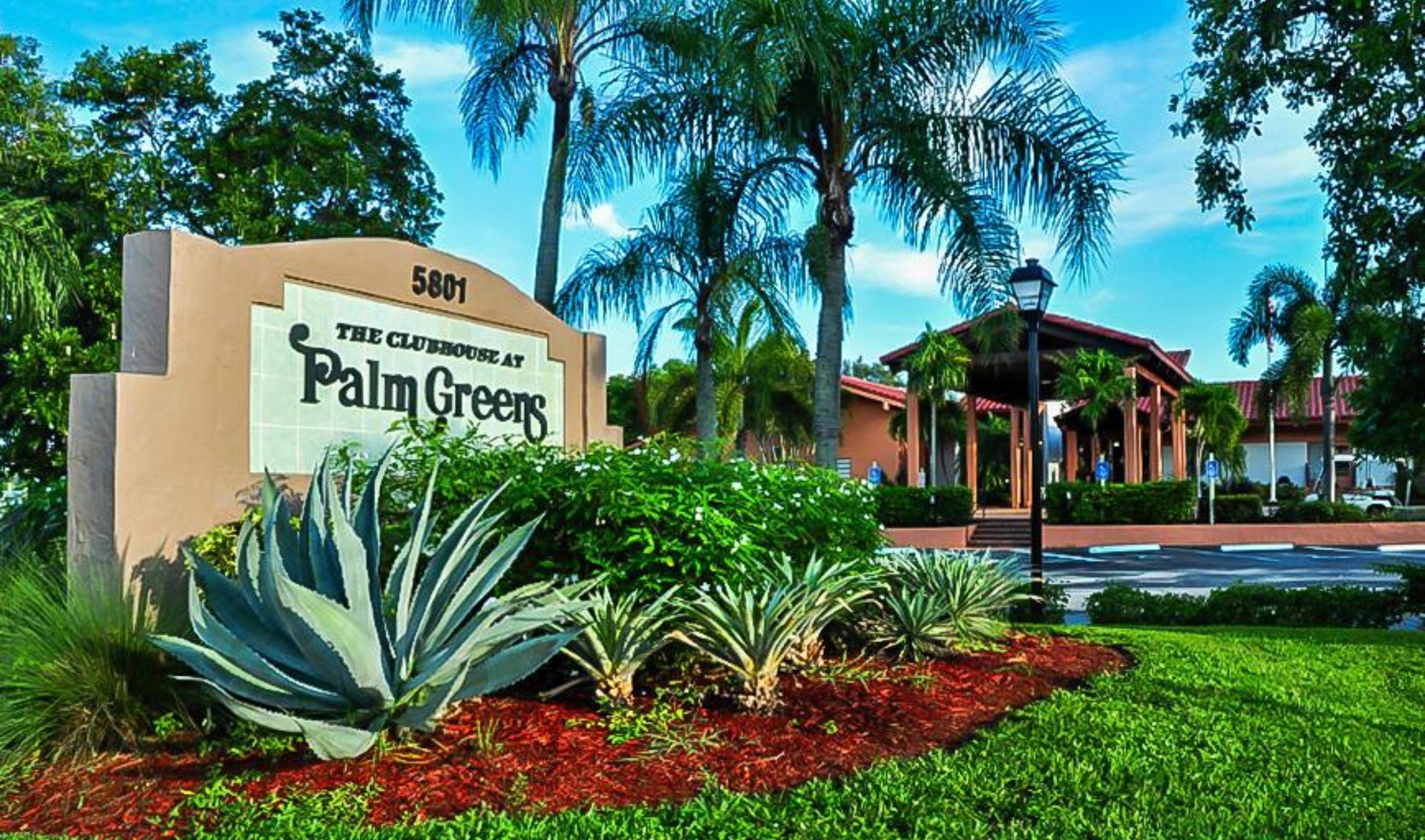 Palm Greens clubhouse entrance with its community sign, palm trees, and landscaped garden.