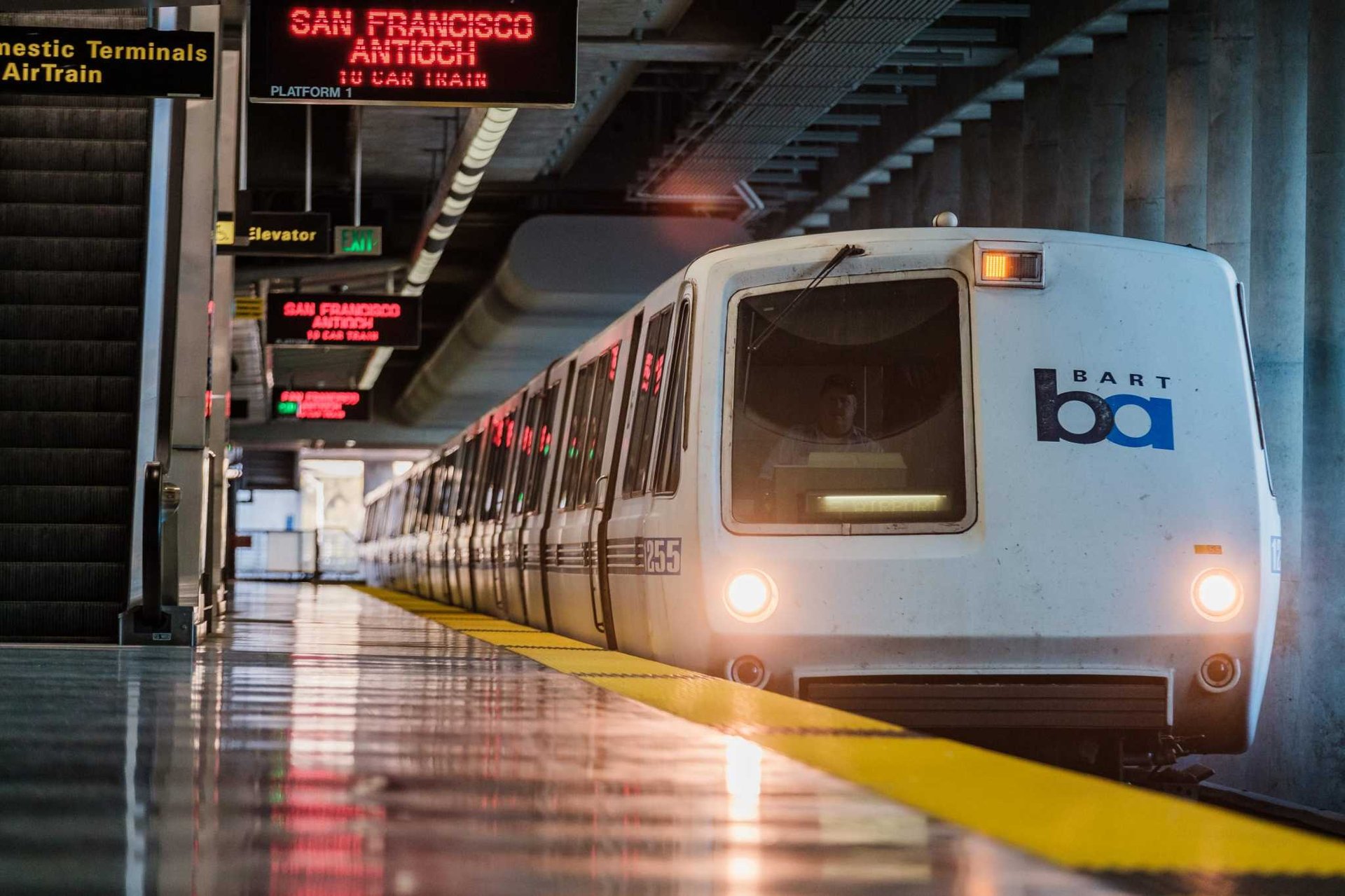 A Bay Area Rapid Transit (BART) train in San Francisco, featuring a sleek design with a white and blue exterior. The train is stationed at a platform, with open doors ready for passengers to board. Overhead signage displays the train's destination, and the platform is equipped with safety markings and lighting.