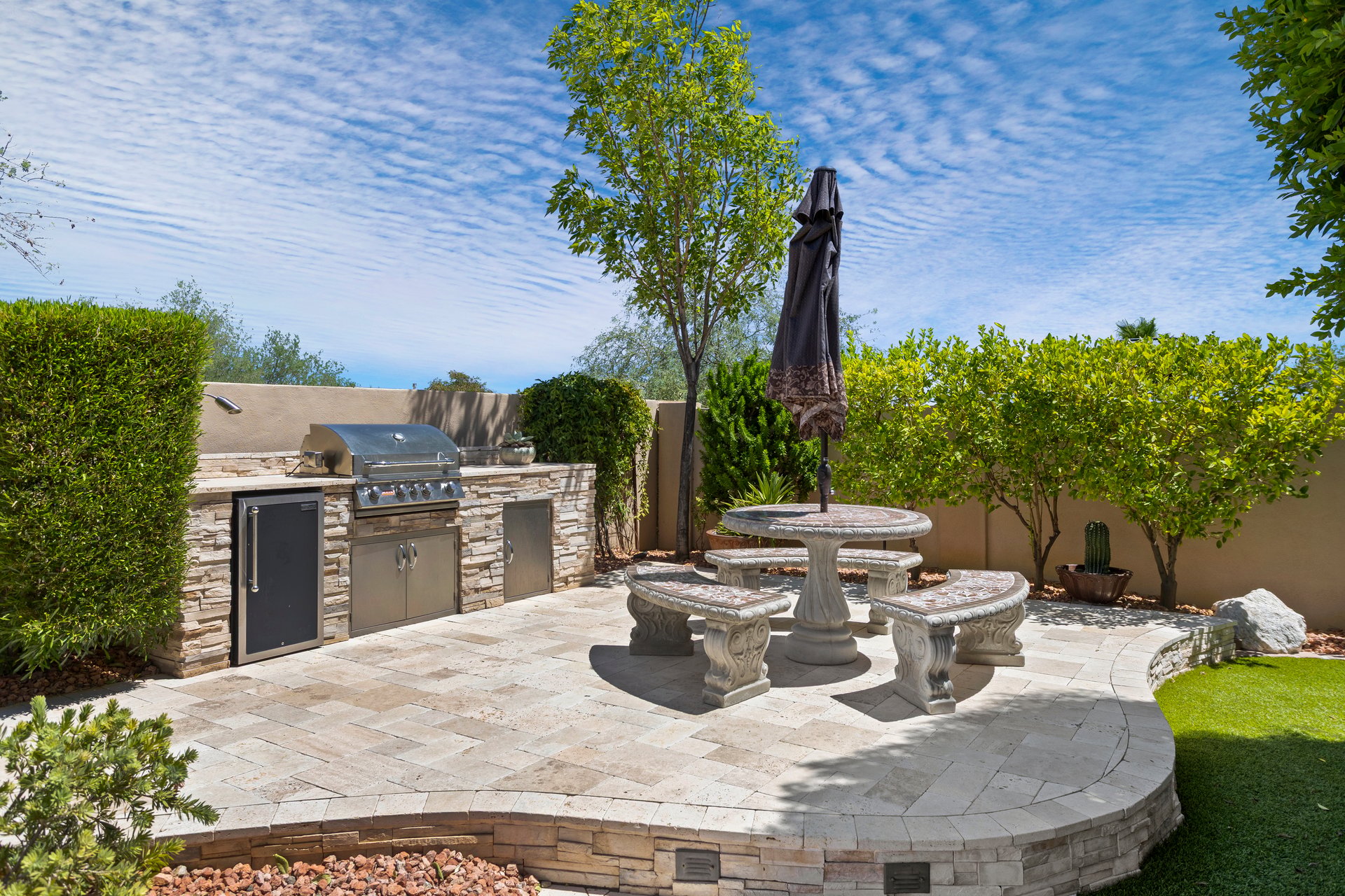 A landscaped backyard patio with a built-in grill and mini fridge, stone dining table with curved benches, and an umbrella under a bright blue sky.