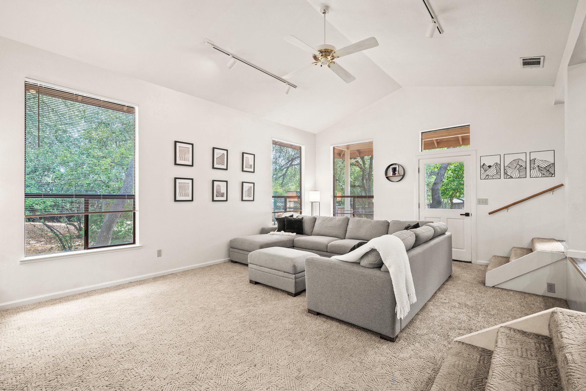 Furnished living room with a gray sectional, tall windows overlooking trees, and a staircase.