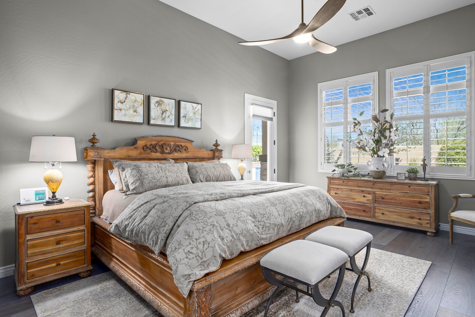 A furnished bedroom with a carved wooden bed, matching nightstands and dresser, gray walls, large windows with shutters, and a ceiling fan.