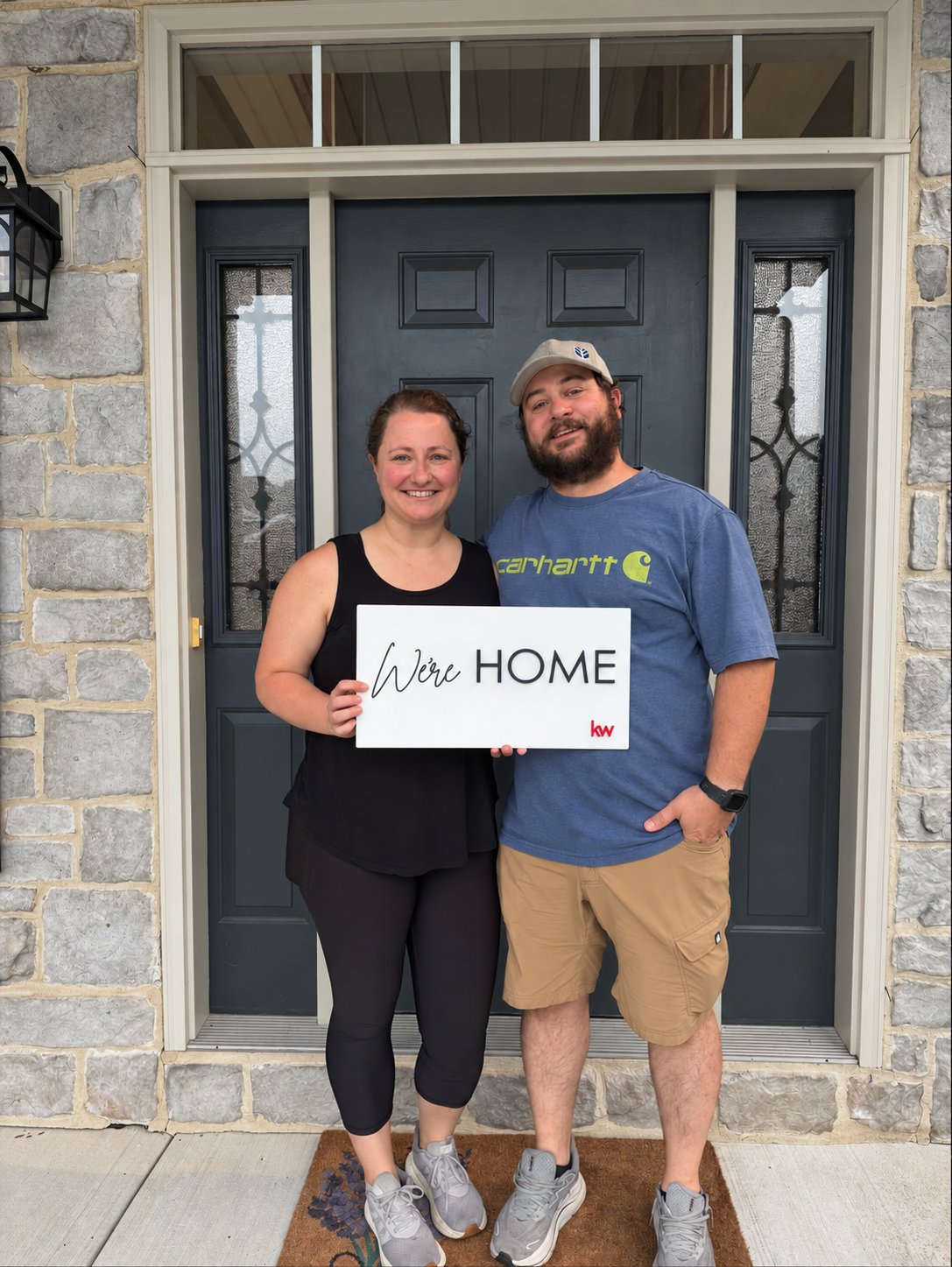 Shelby and Jordan celebrating at the front door of their new home with a We’re Home sign