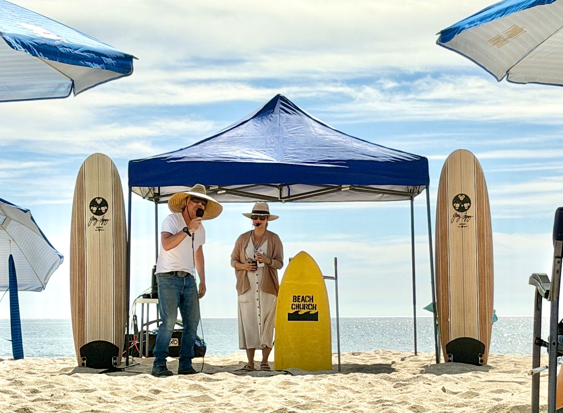 Two speakers beneath a blue canopy beside Beach Church signs and the ocean.