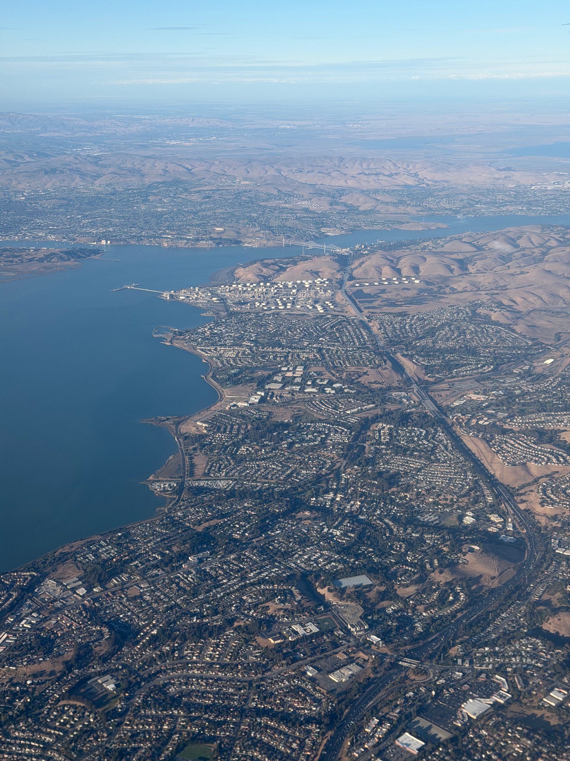 Aerial view looking north along the East Bay shoreline, with bayside neighbourhoods and freeway on the left and dry inland hills rising to the right.