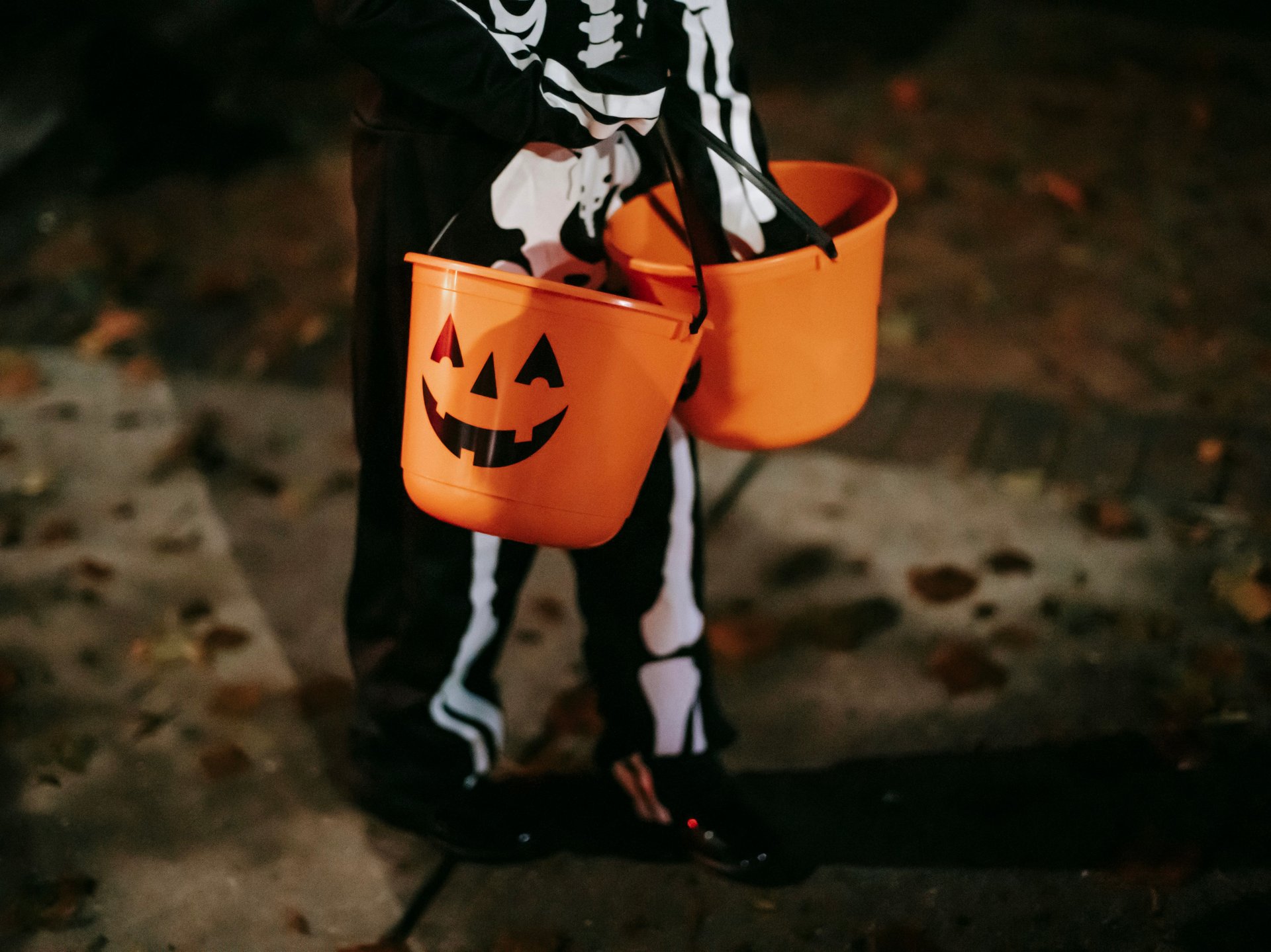 Child in a skeleton costume carrying two orange jack-o’-lantern trick-or-treat buckets at night.