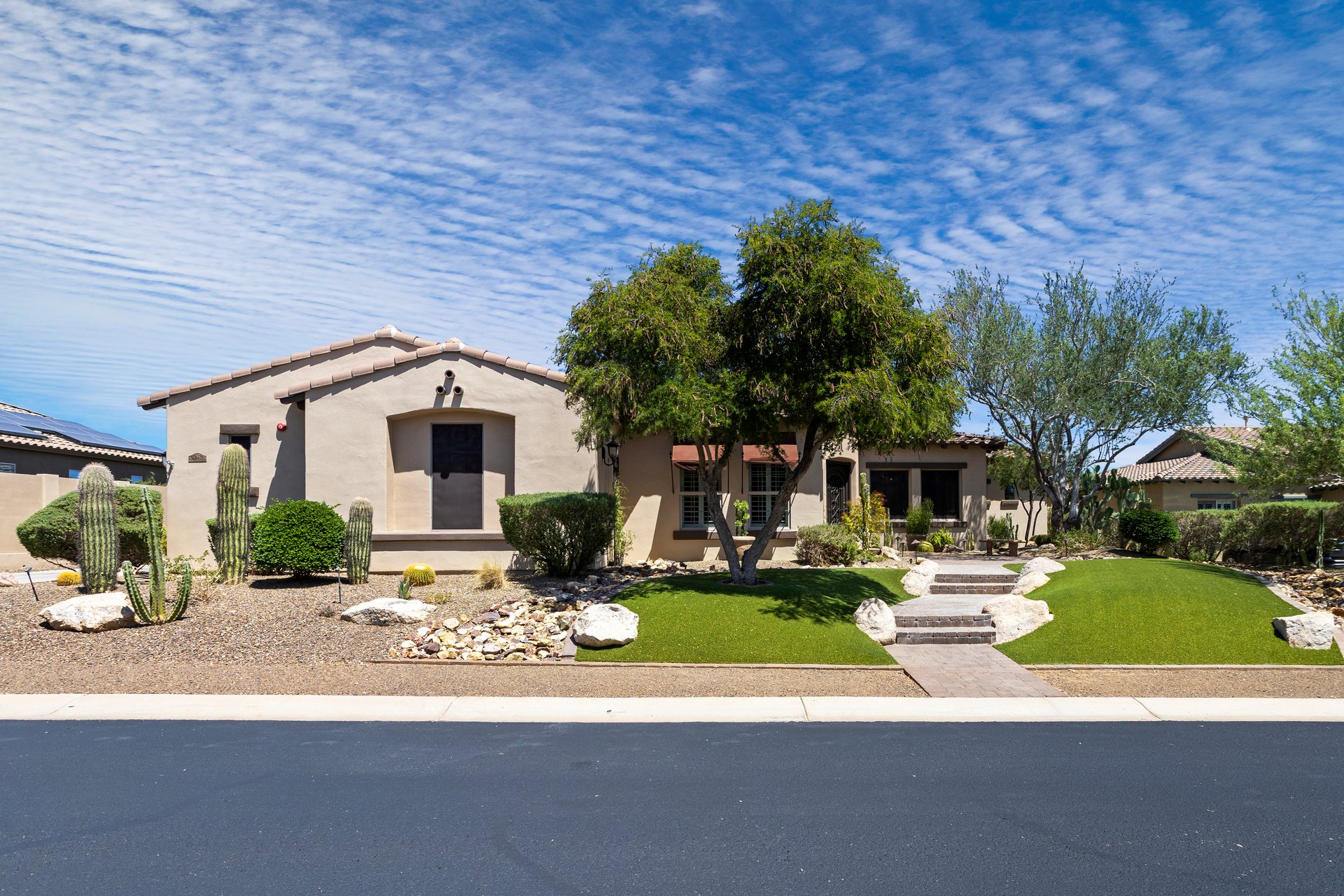 Front exterior of a stucco house with desert landscaping, cacti, trees, and a walkway leading to the entry under a blue sky.
