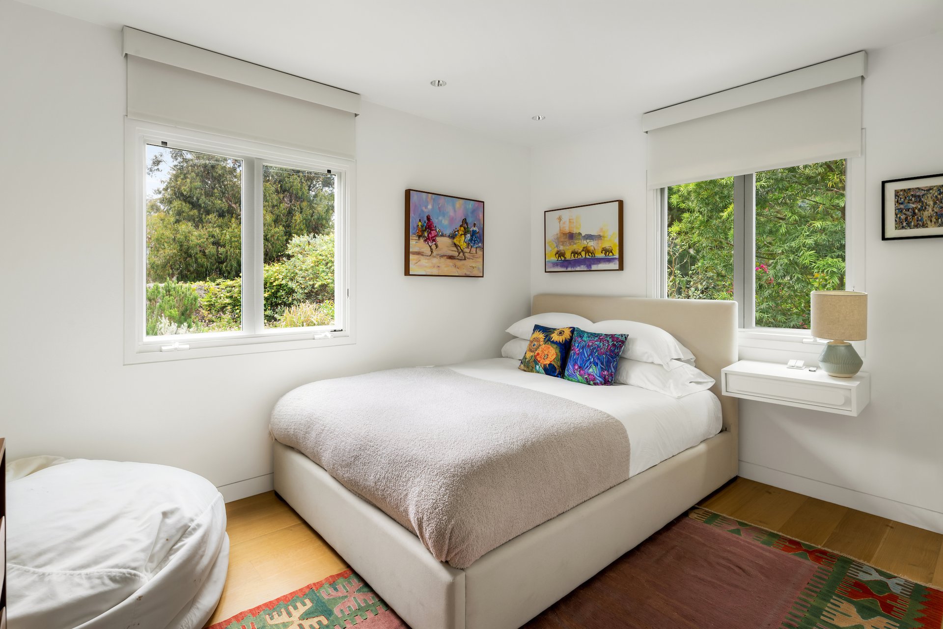 Bright bedroom with a white upholstered bed, colorful pillows, and large windows overlooking greenery.