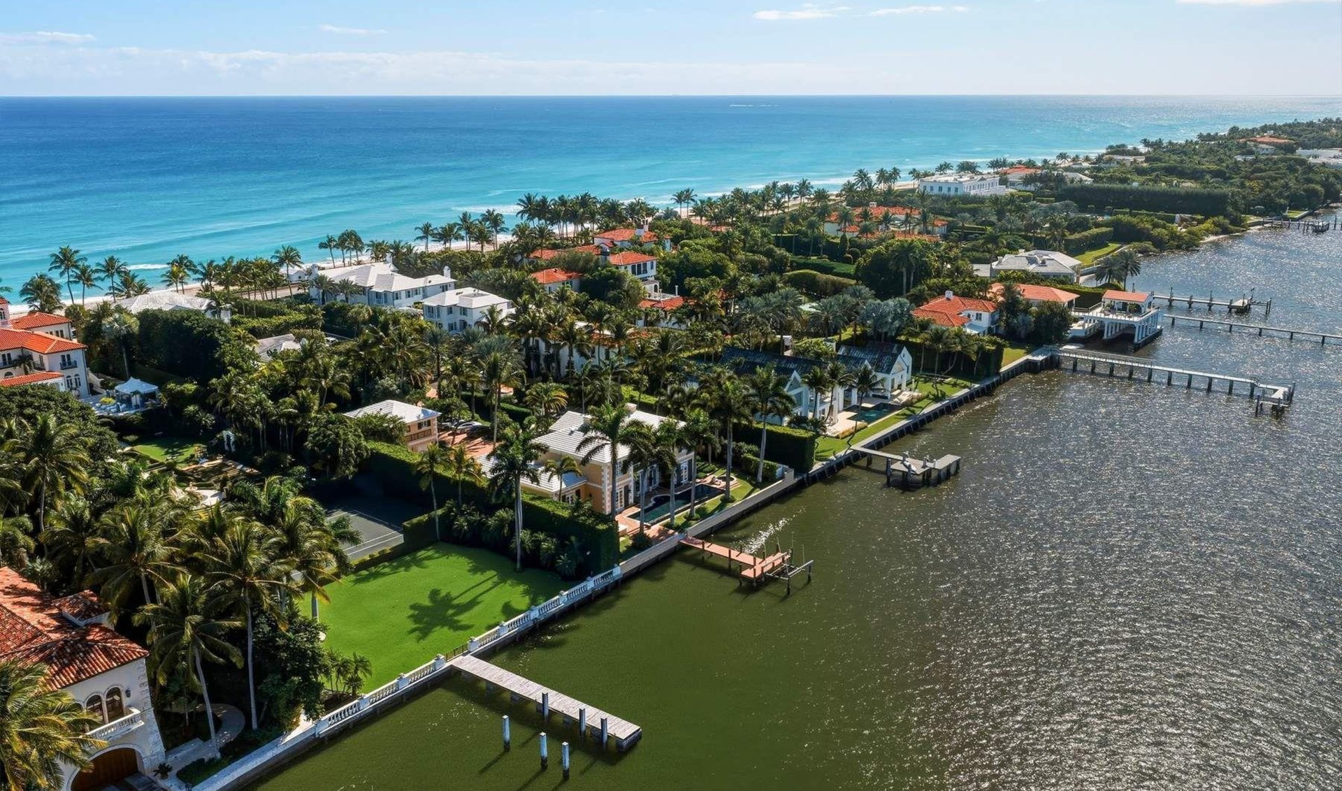 Aerial view of waterfront homes and docks between a palm-lined beach and the ocean.