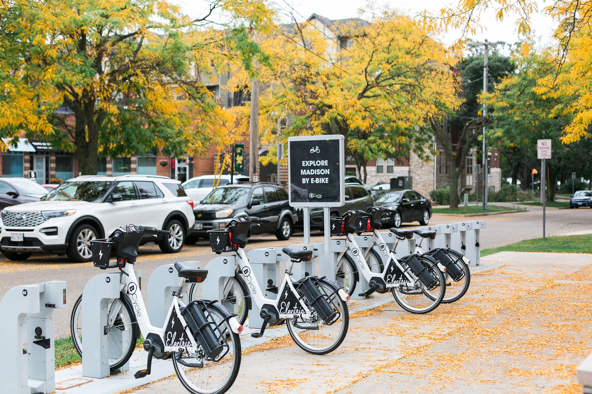 A row of white Madison BCycle electric bikes at a docking station on a tree-lined street in fall, highlighting the bike-sharing infrastructure that supports the most walkable neighborhoods in Madison, Wisconsin.