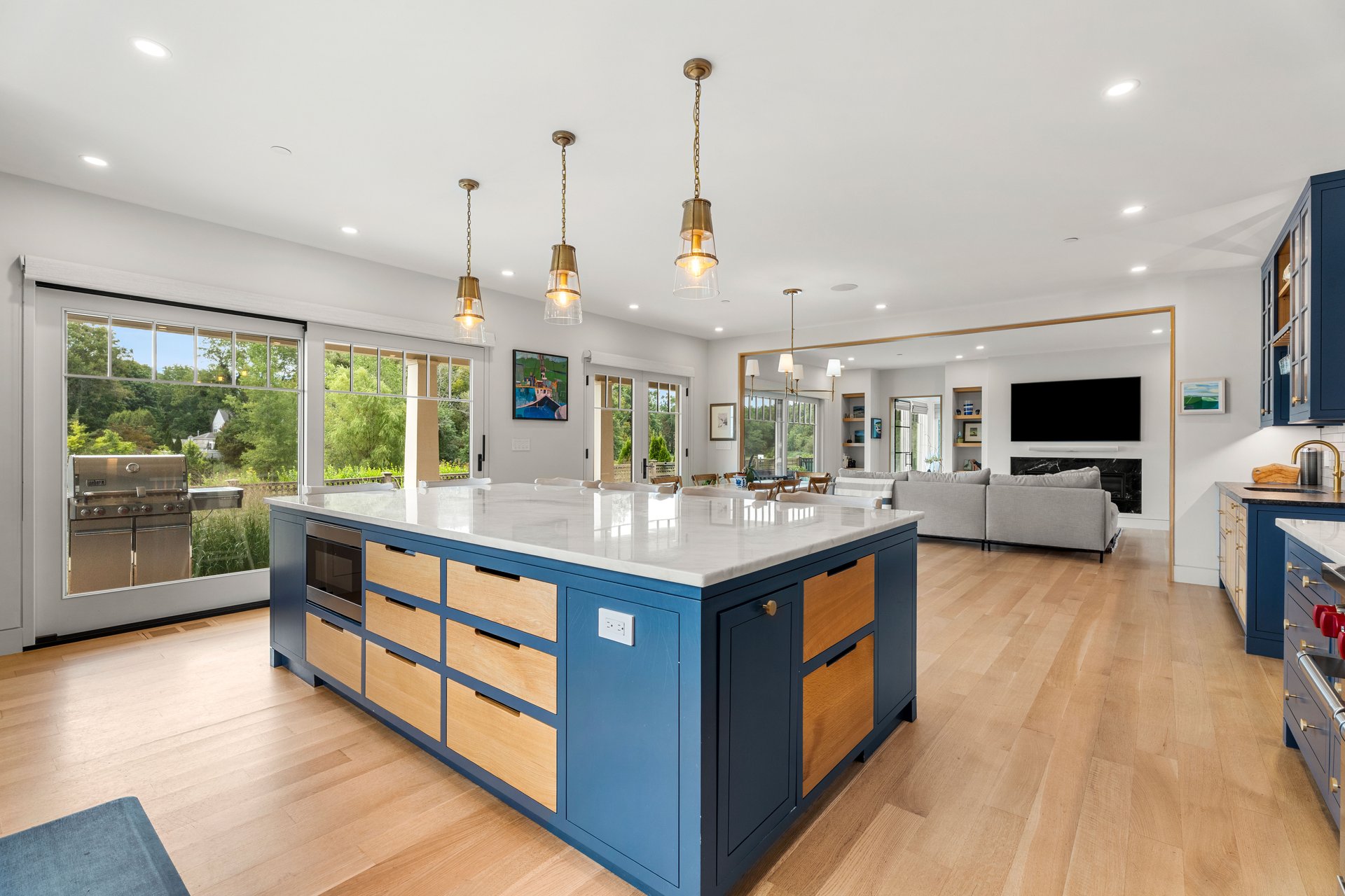 Open-plan kitchen with a blue-and-wood island, brass pendants, and large windows overlooking greenery.