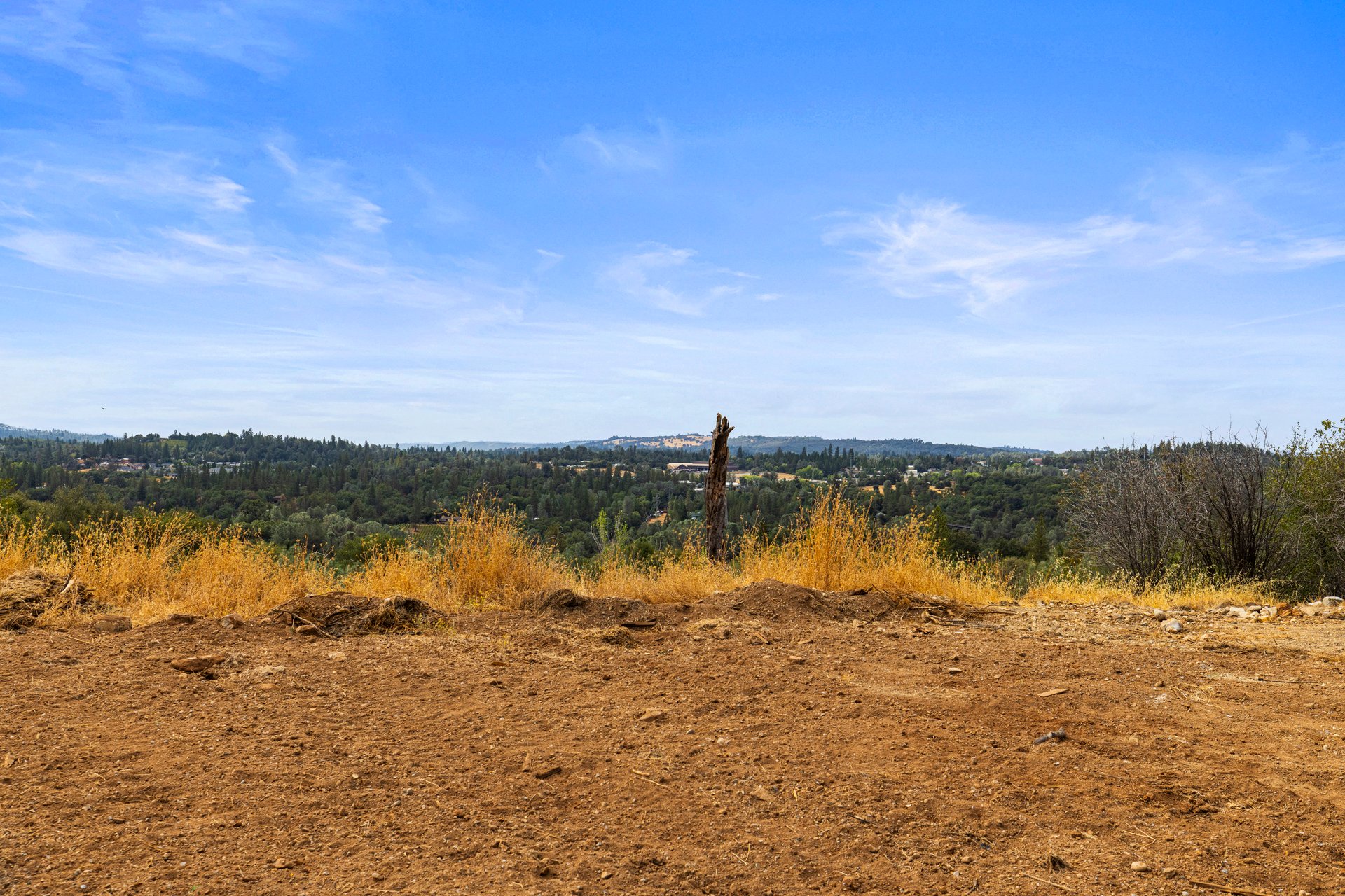 Cleared dirt lot overlooking wooded rolling hills, with golden grasses and a tree stump.
