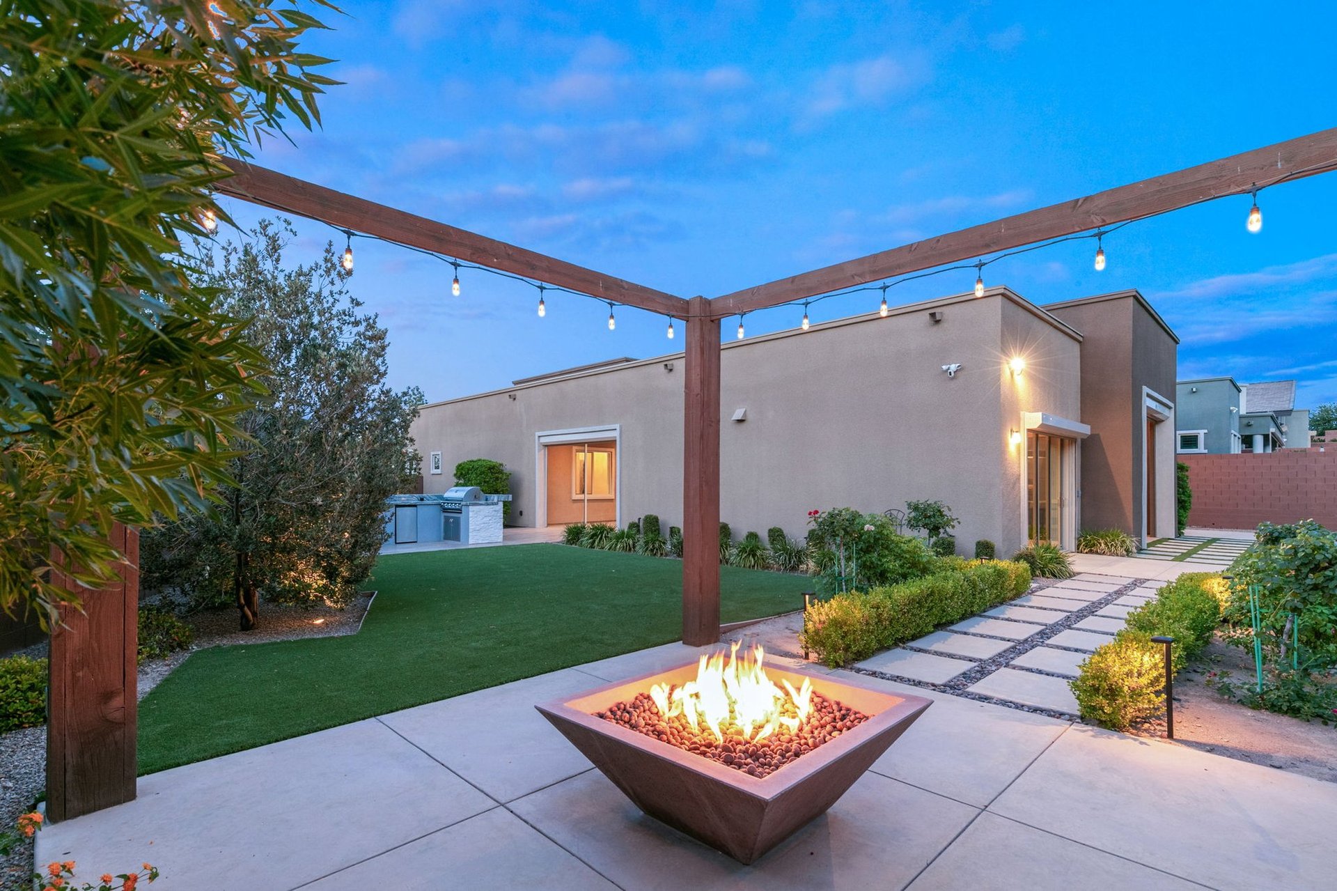 Twilight view of a modern Summerlin luxury home backyard with a glowing fire pit, wood pergola with string lights, lawn and desert landscaping in Las Vegas