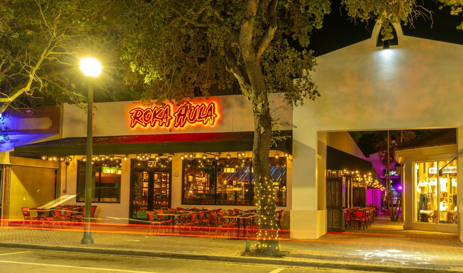 Roka Hula restaurant exterior at night, with its glowing sign and outdoor dining tables.