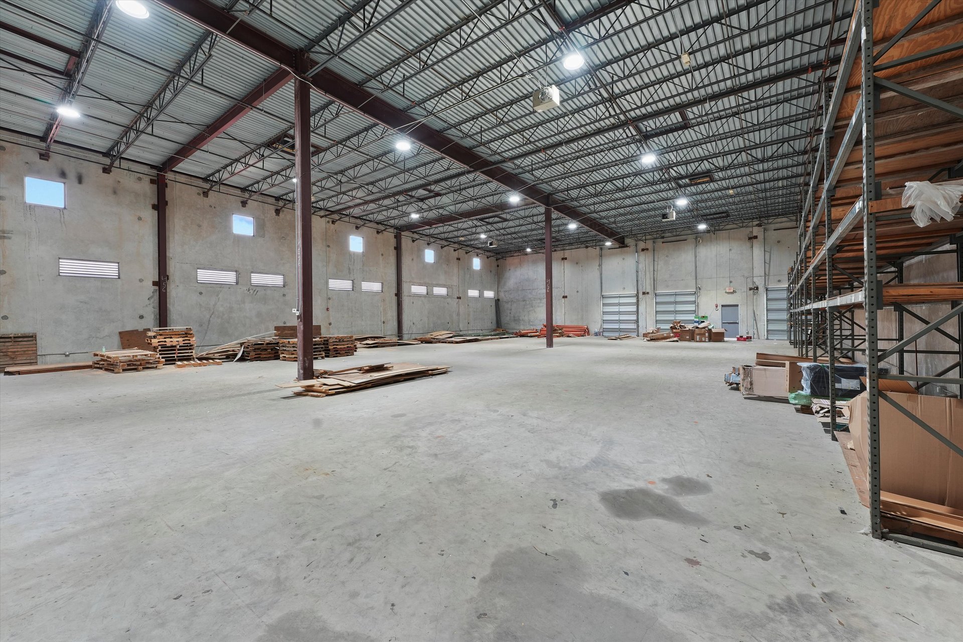 Large warehouse interior with high metal-truss ceiling, open concrete floor, loading doors, and perimeter storage racks.
