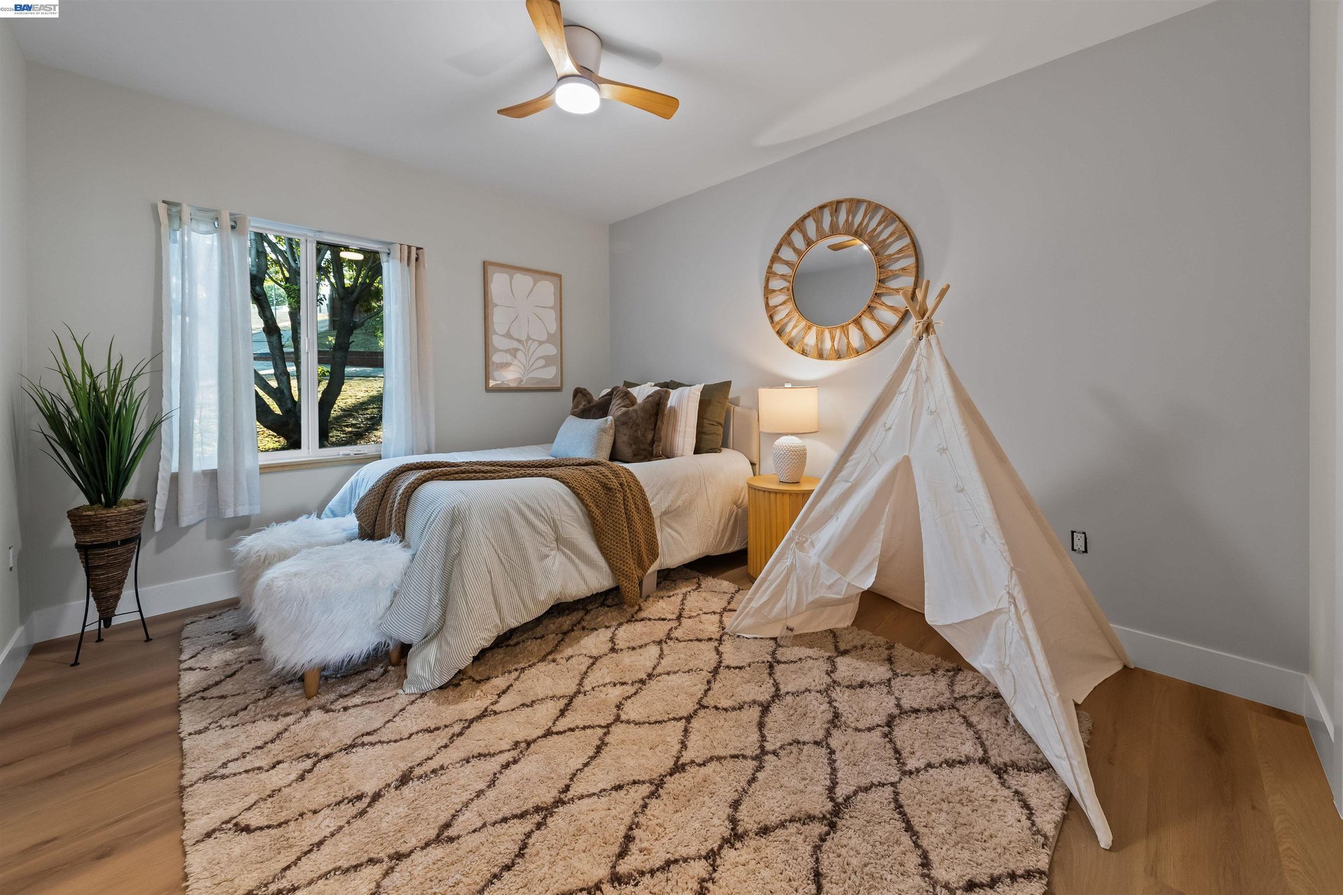 Staged bedroom with a white bed, children’s teepee, sunburst mirror, and large patterned rug.