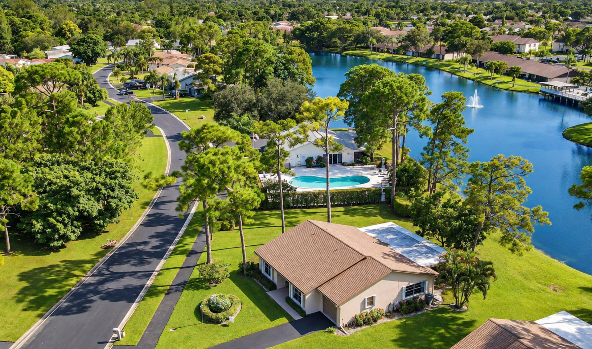 Aerial view of a leafy waterfront neighborhood with a curving road, lake, and homes with pools.