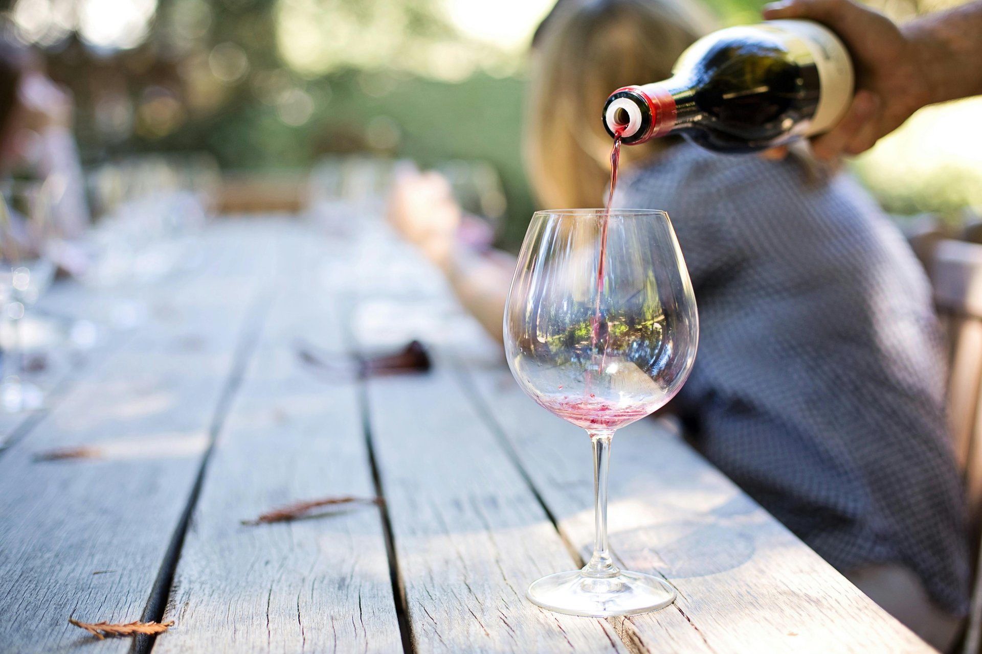 A glass is being filled with red wine from a bottle, set on a wooden table with blurred background elements.
