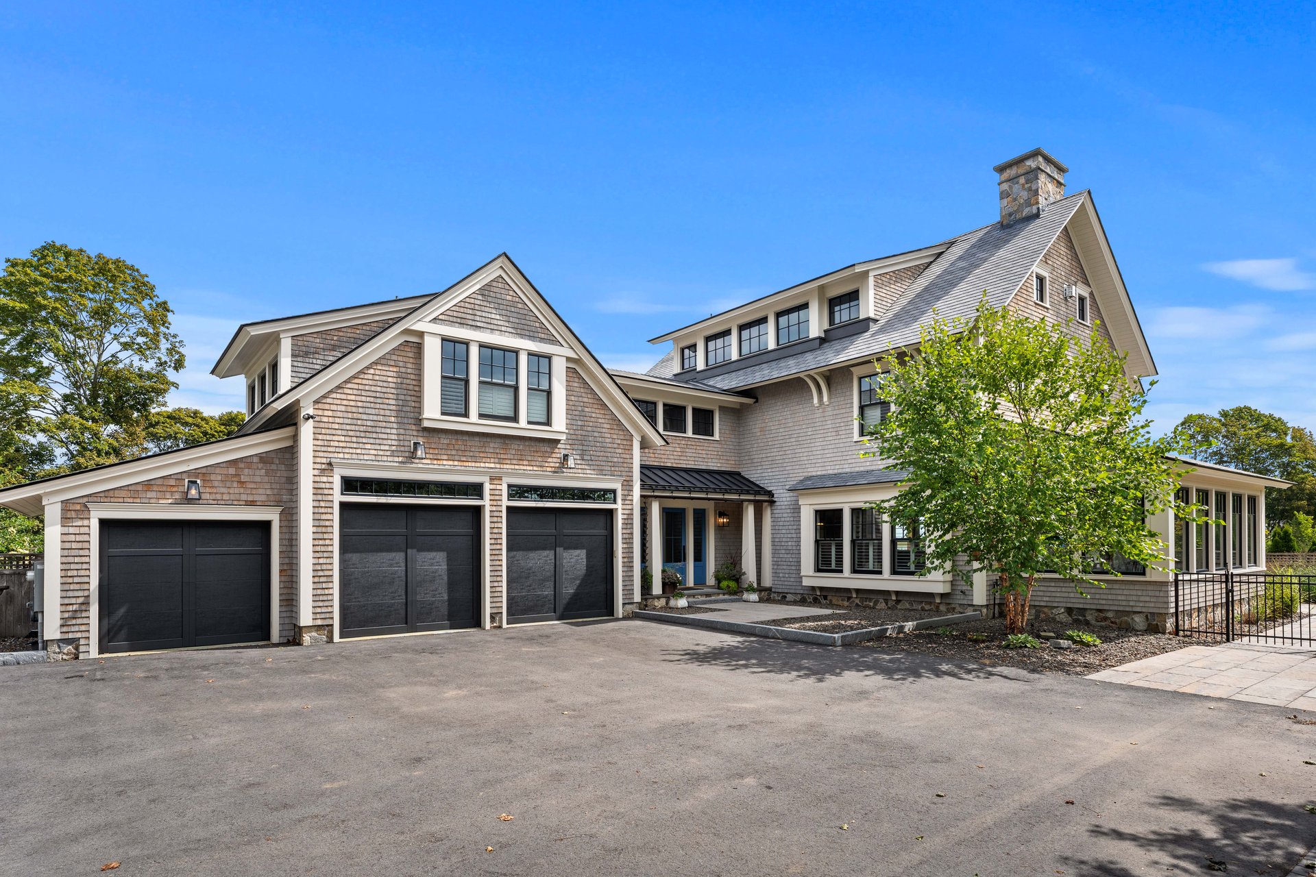 Shingle-style home with three dark garage doors, multiple gables, and a stone chimney.