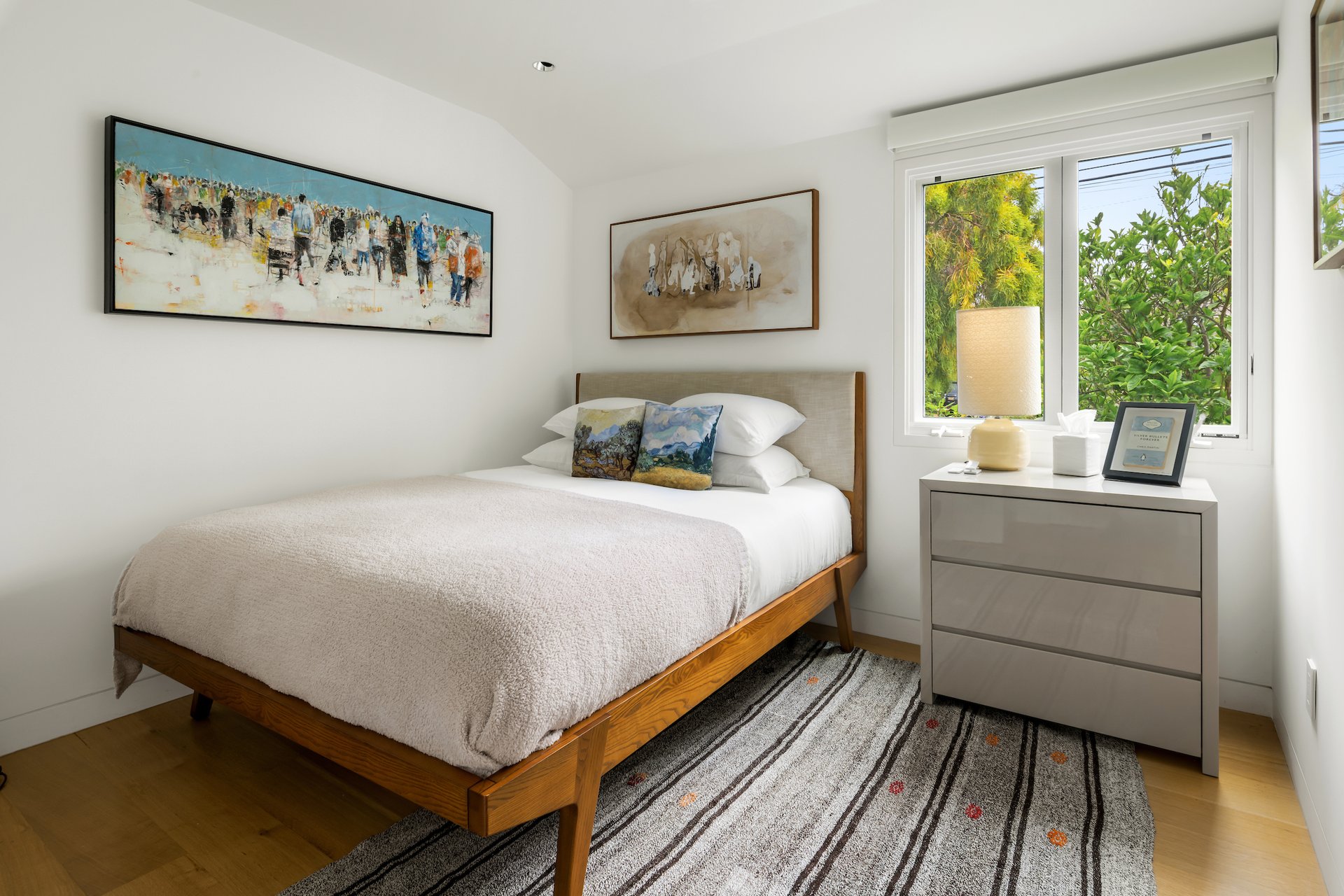 Bright bedroom with a wood-framed bed, abstract wall art, and a window overlooking greenery.